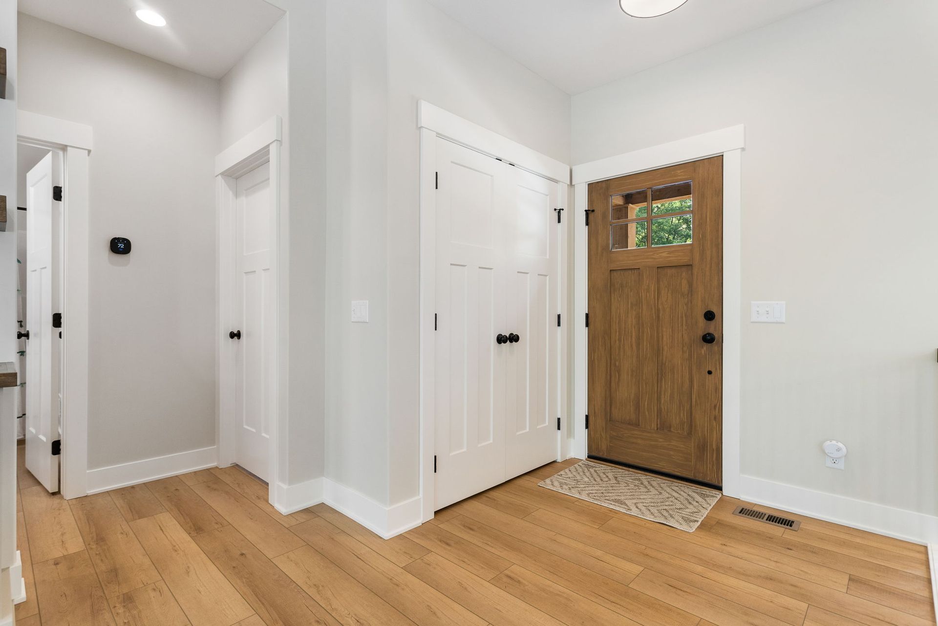 A bright indoor entryway featuring light oak wood floors, white doors, and a natural wood-toned front door.