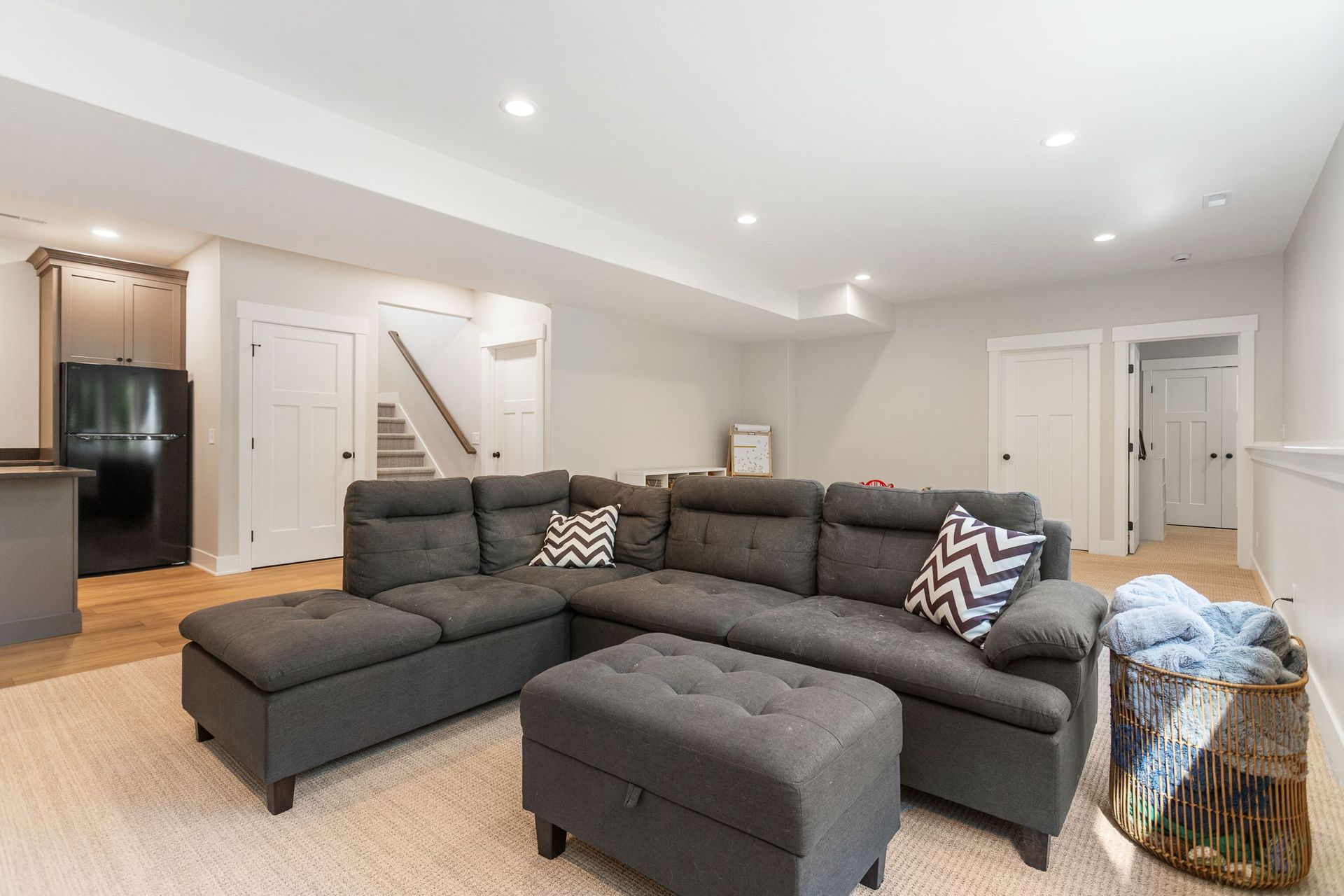 A spacious basement living area features a grey sectional sofa, an ottoman, a kitchen corner, and neutral carpeting.