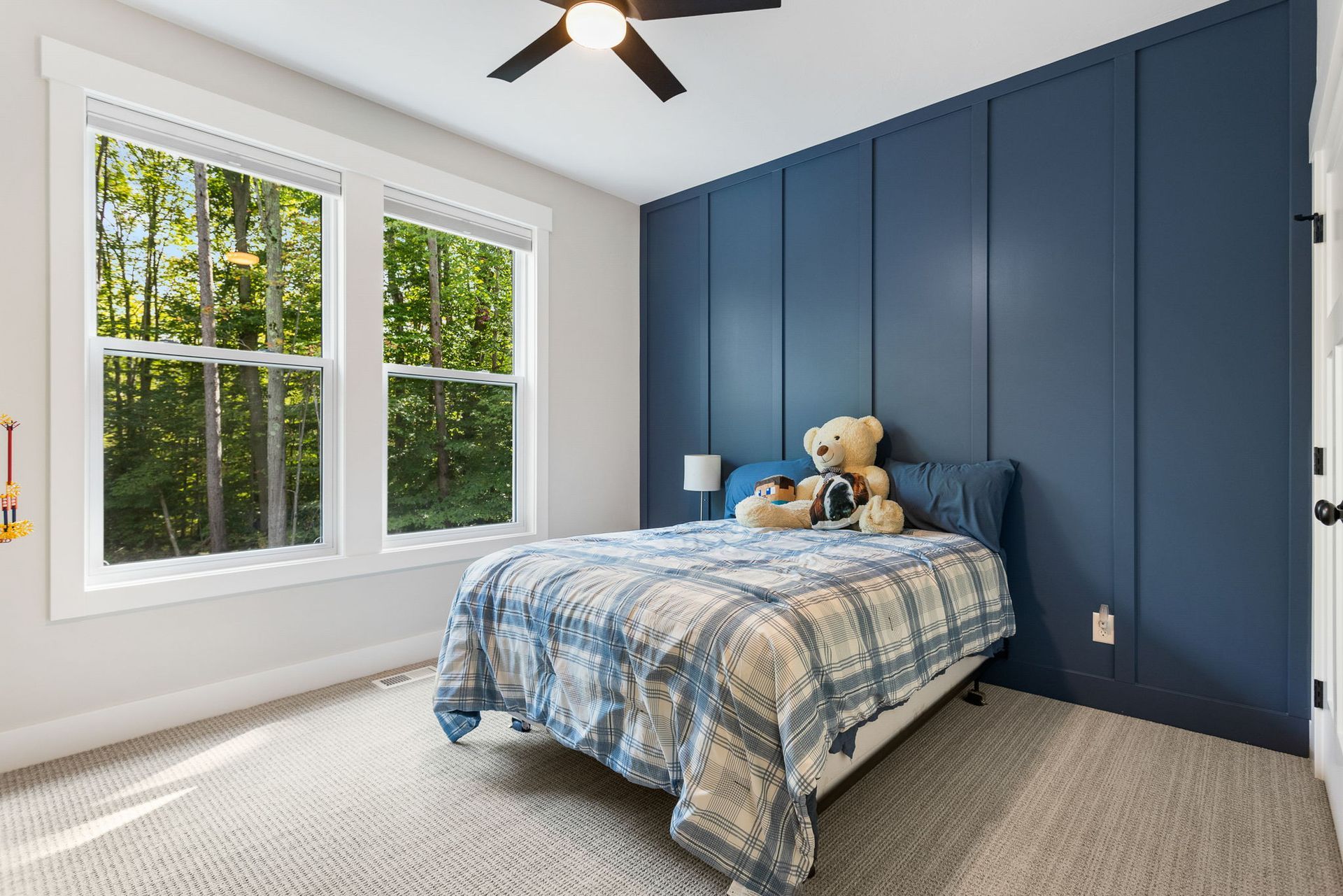 A bedroom with light carpet, white walls, and a deep blue paneled accent wall featuring a bed with a plaid comforter.