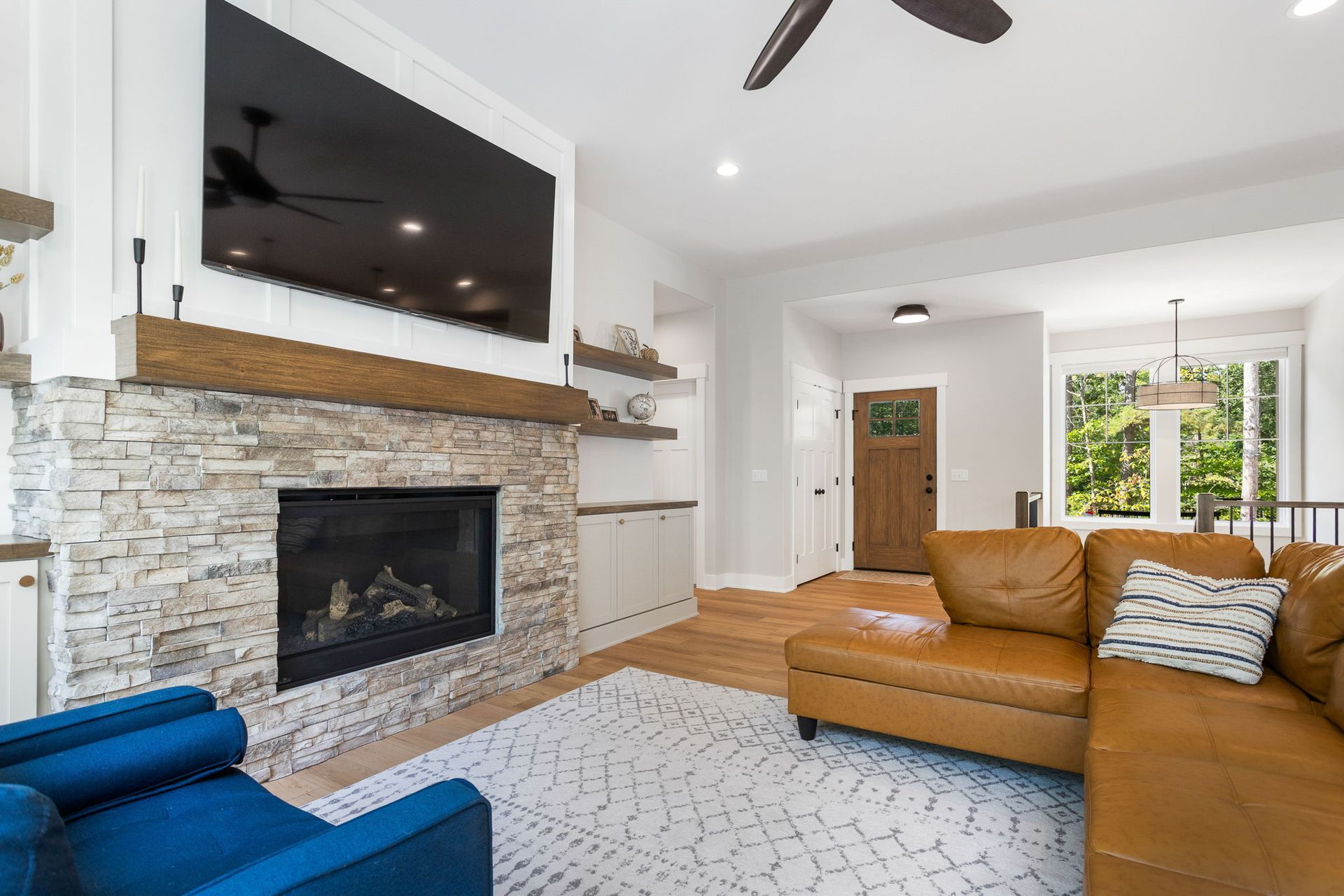 Living room with a stone fireplace, TV, brown leather sectional, blue armchair, and light hardwood floors.