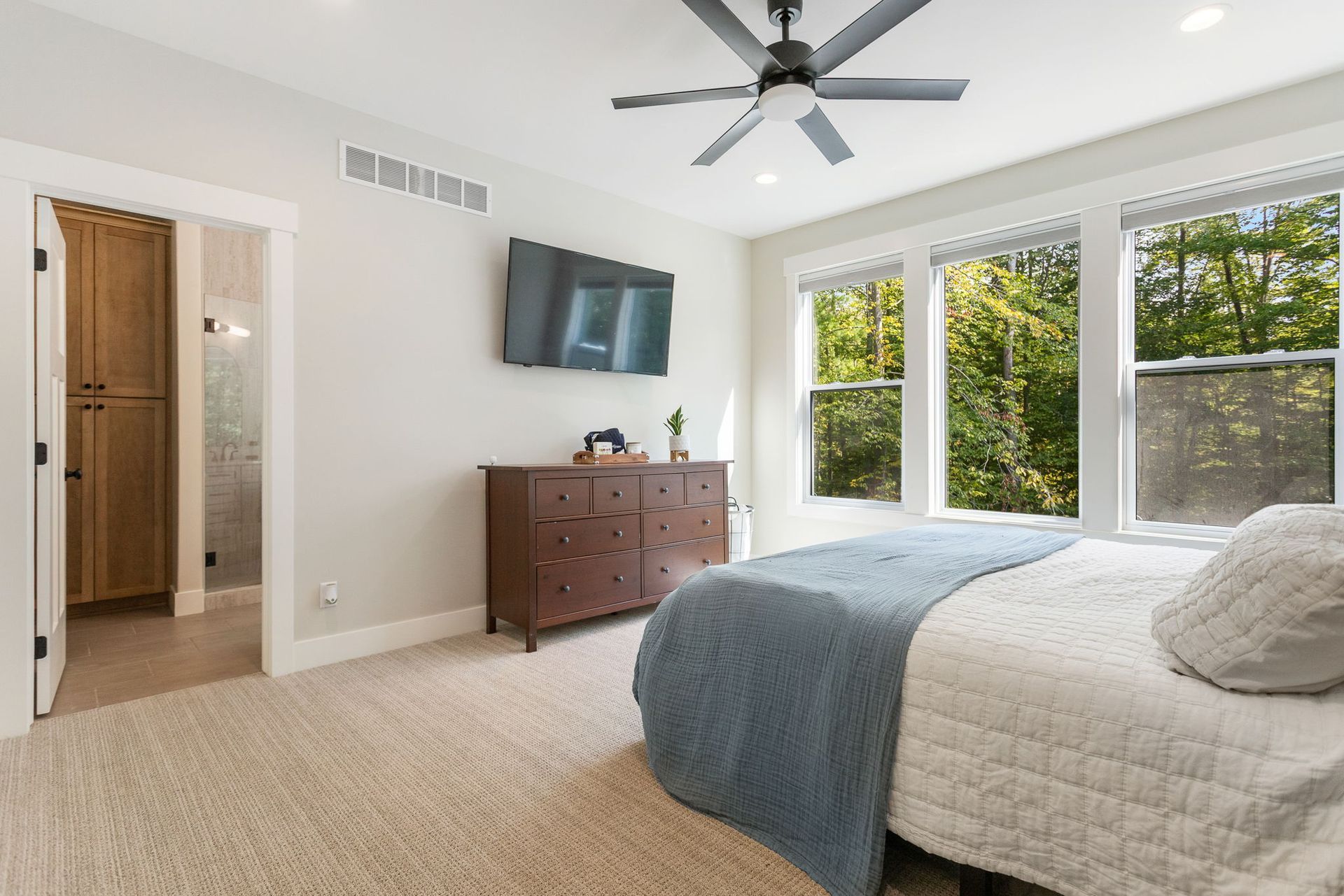 A bright bedroom with a white bed, dark wood dresser, wall-mounted TV, and a door leading to a bathroom.