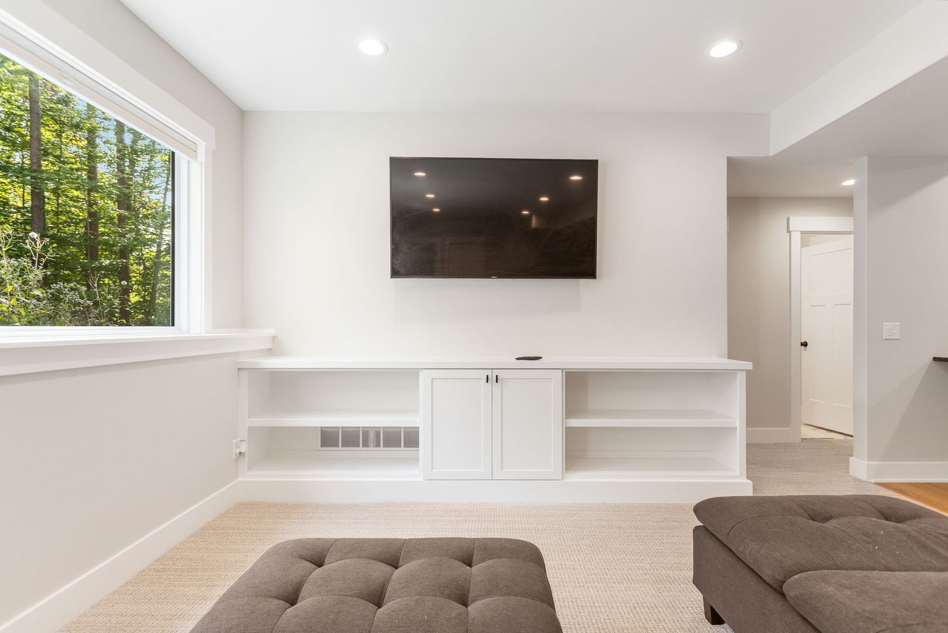 A living area with a wall-mounted TV above a white built-in media console, flanked by windows and light carpeting.
