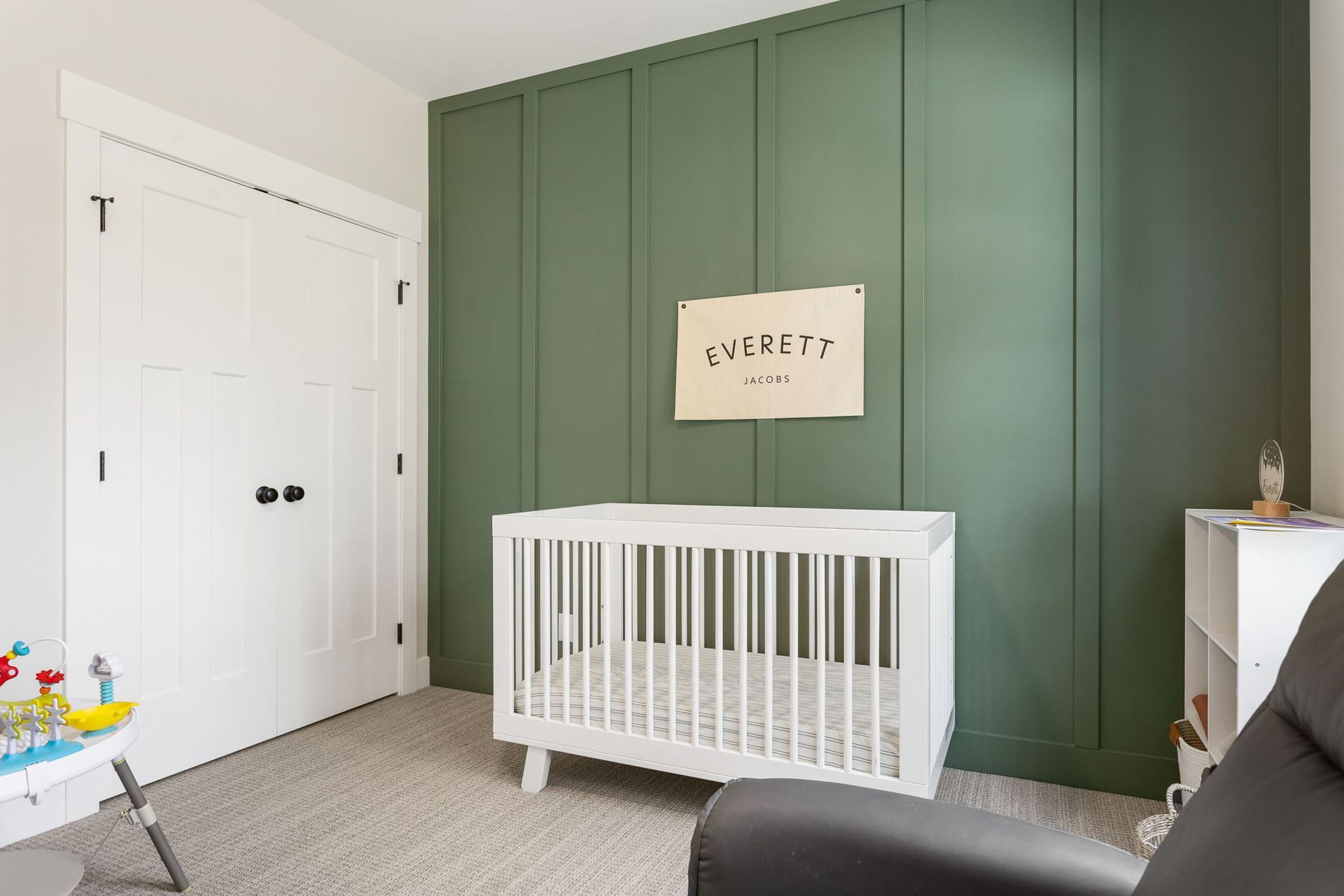 A white crib sits in front of a green board-and-batten accent wall in a nursery with white doors and carpet.