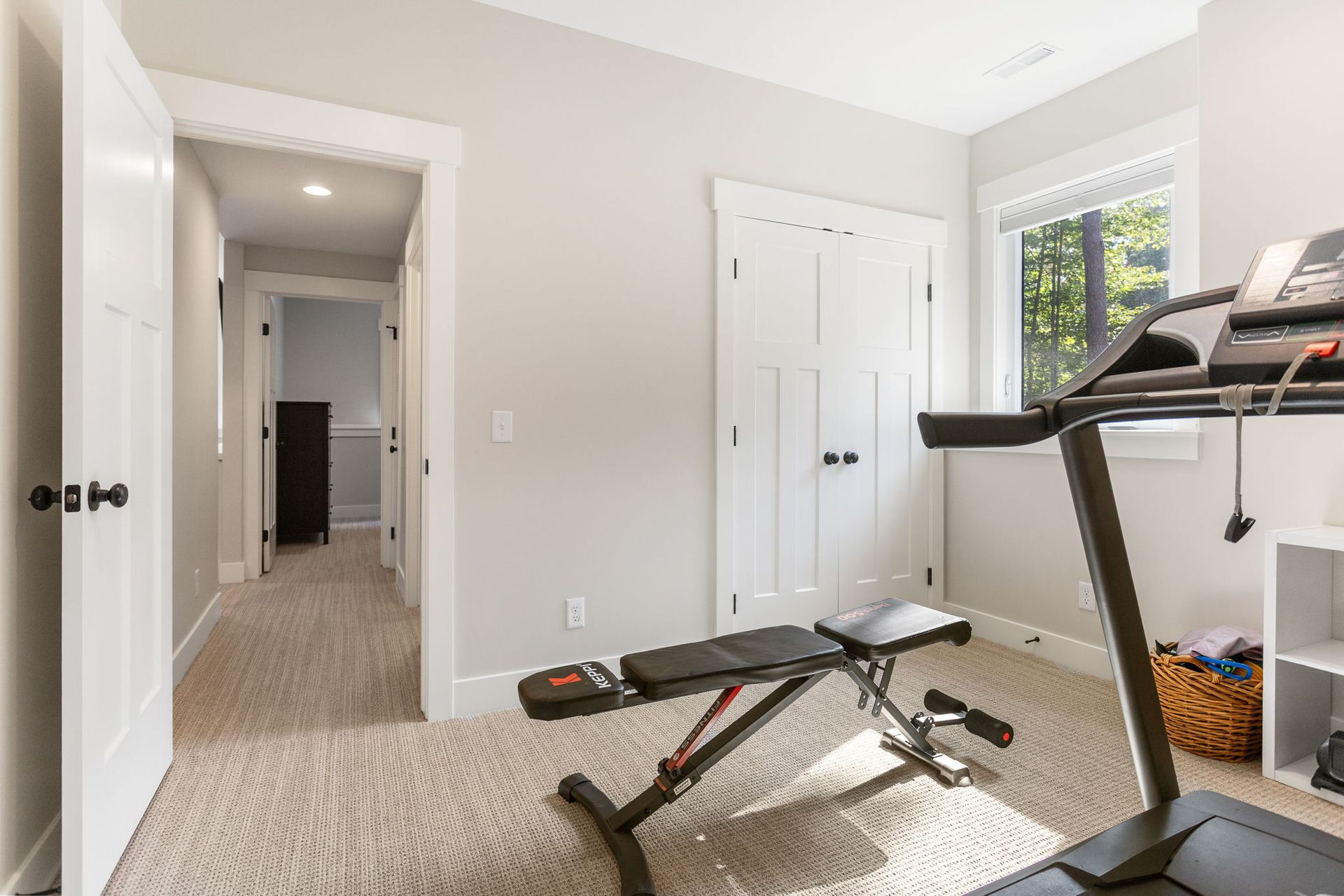 A home gym room features a treadmill and a weight bench on carpeted flooring, with a doorway leading to a hallway.