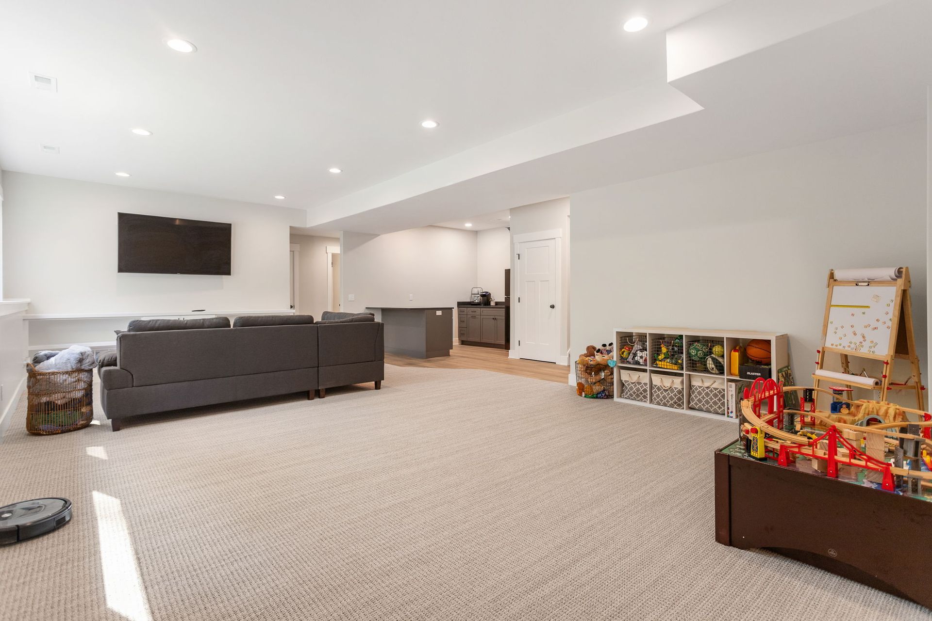 A modern finished basement with a grey sofa, television, open wet bar area, and a children's play area with storage bins.