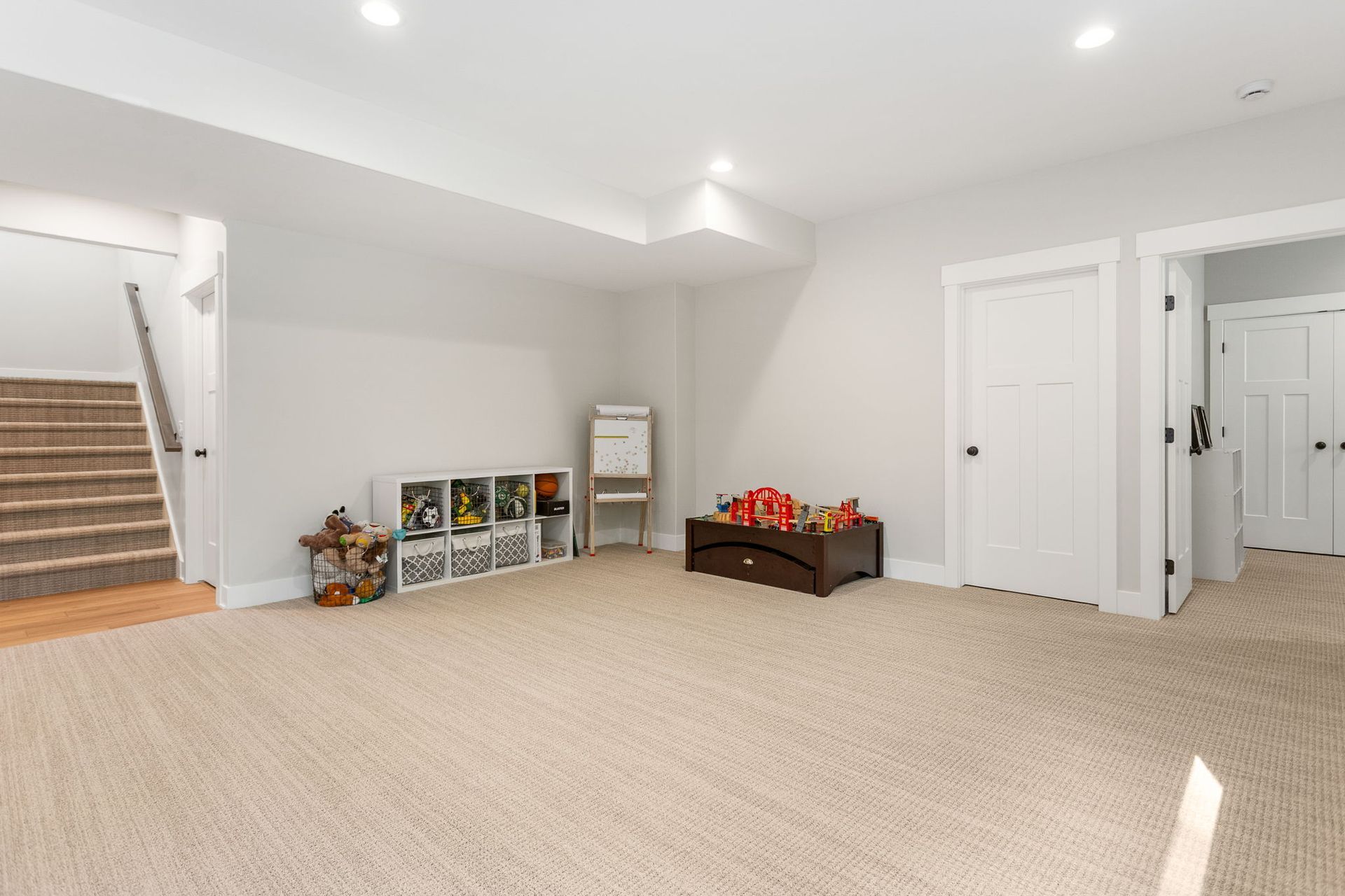 A carpeted basement with a staircase on the left, toy storage, and two white doors on the right.
