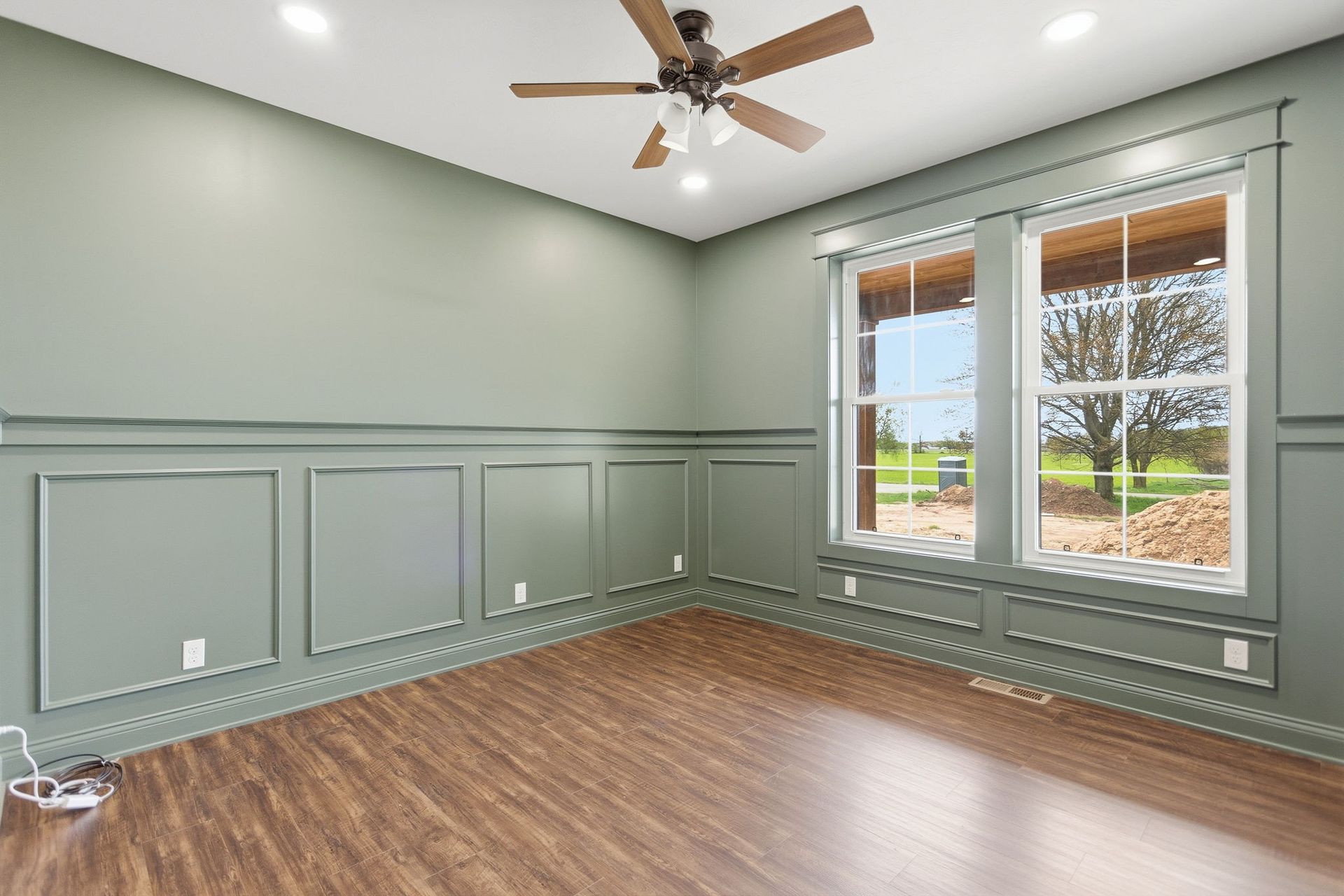 Empty room with sage green wainscoting, wood floor, ceiling fan, and large windows with blinds