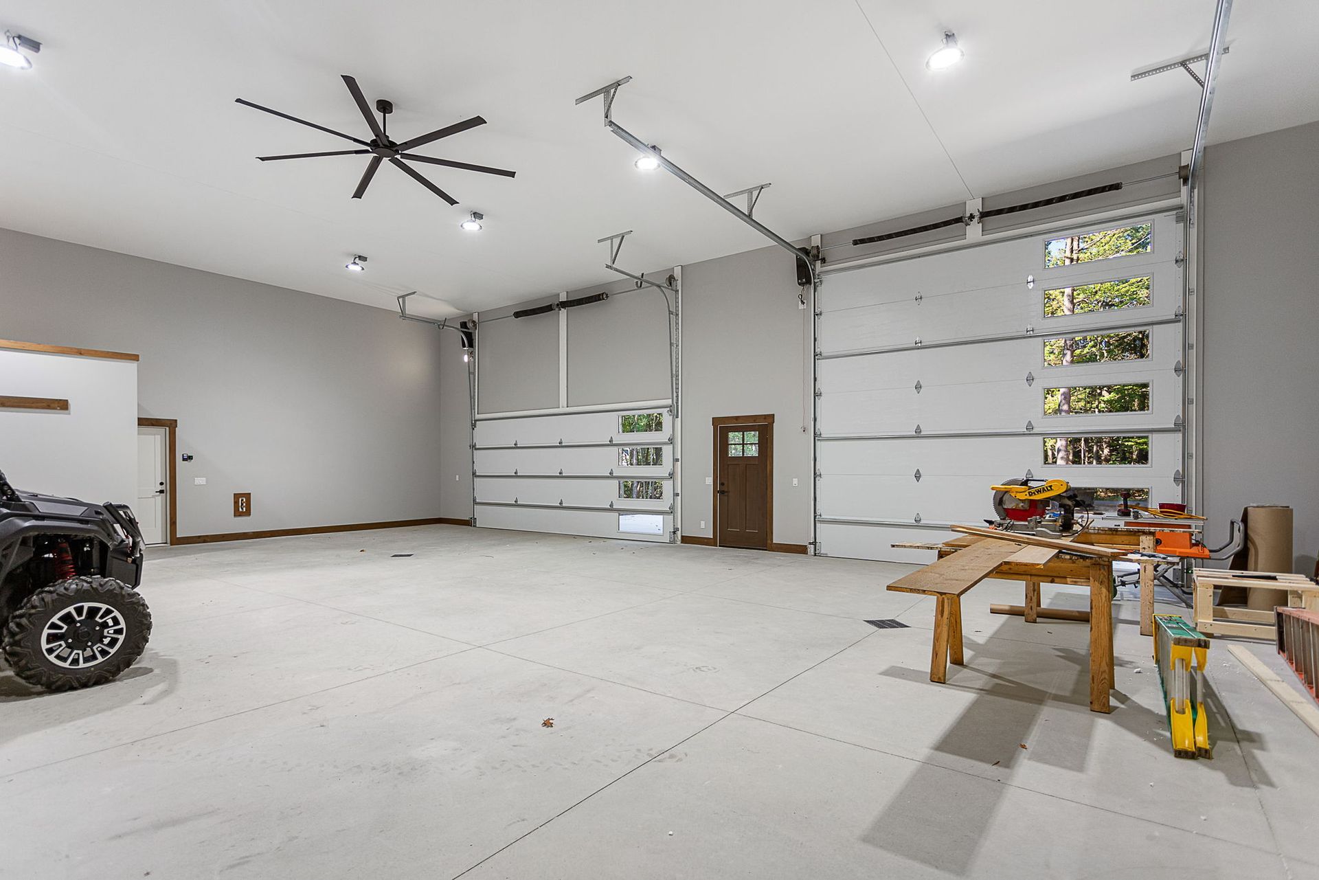 A spacious, well-lit modern garage with gray walls, a ceiling fan, two large overhead doors, and a wooden workbench.