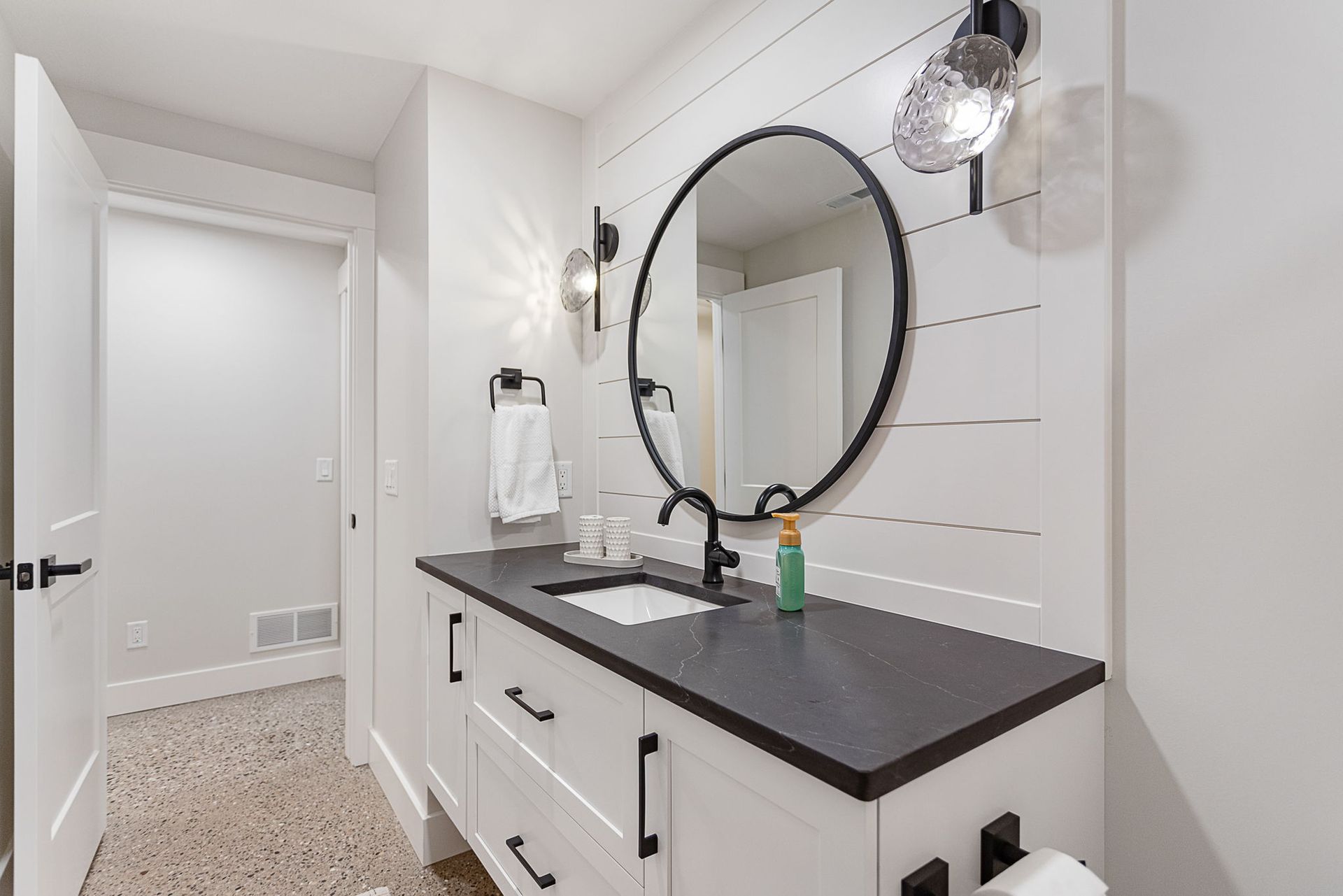 Modern bathroom with a white shiplap wall, round mirror, dark countertop, white vanity, and speckled tile floor.