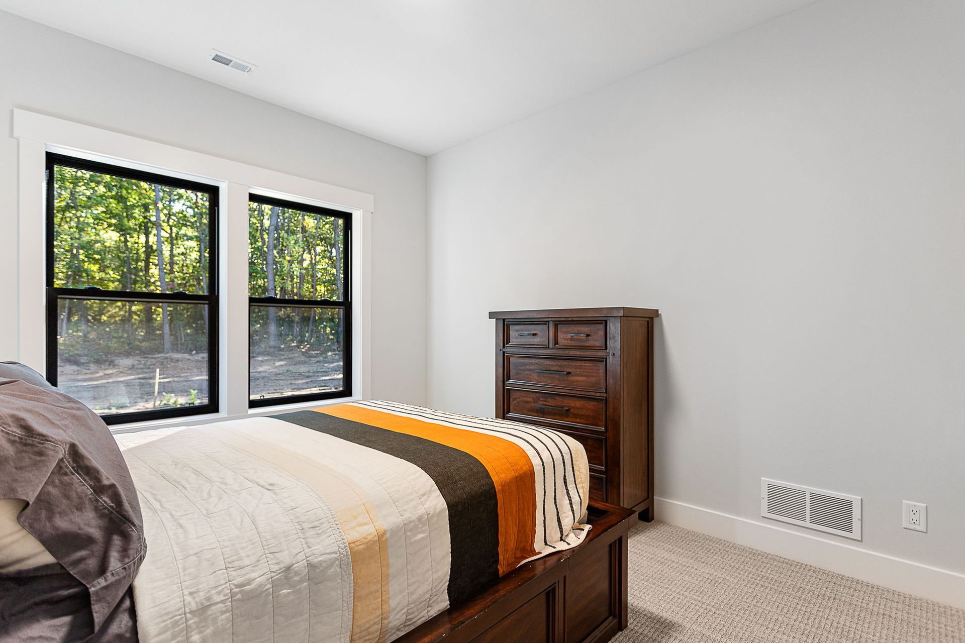 A bedroom with a bed with an orange and gray blanket, a dark wood dresser, and a window overlooking a wooded area.
