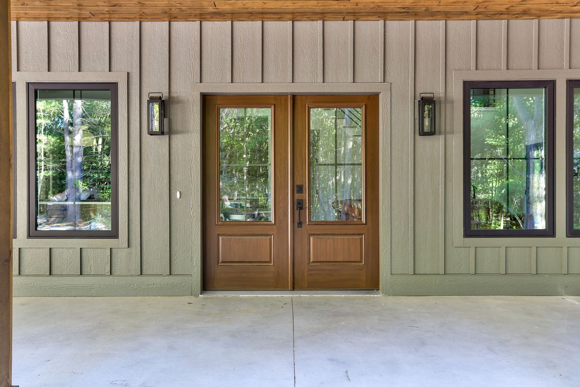 A double wooden door entry with glass panels, flanked by two windows on a board-and-batten wall under a covered porch.