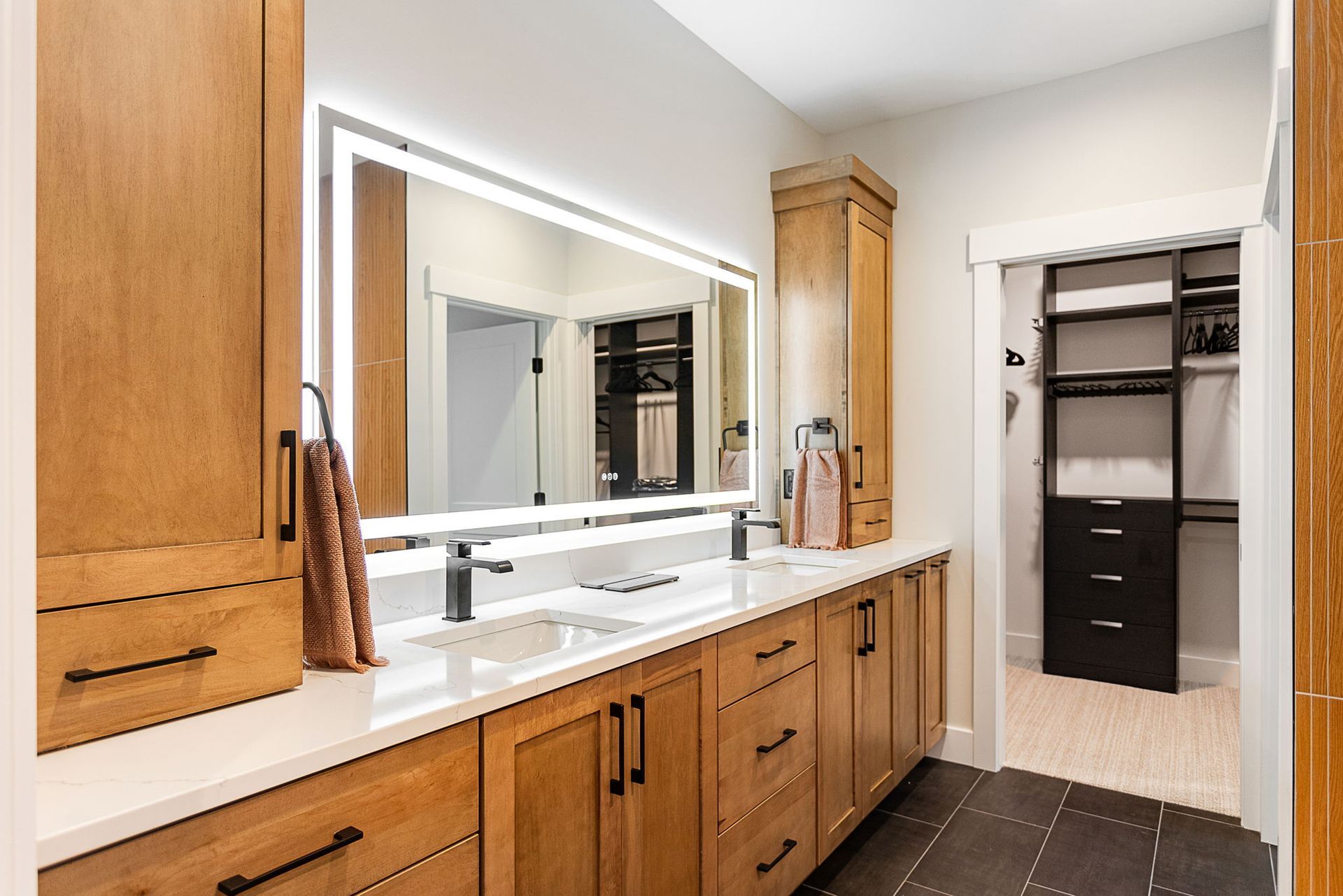 Modern bathroom with light wood cabinets, white countertops, a large LED mirror, and an open walk-in closet.