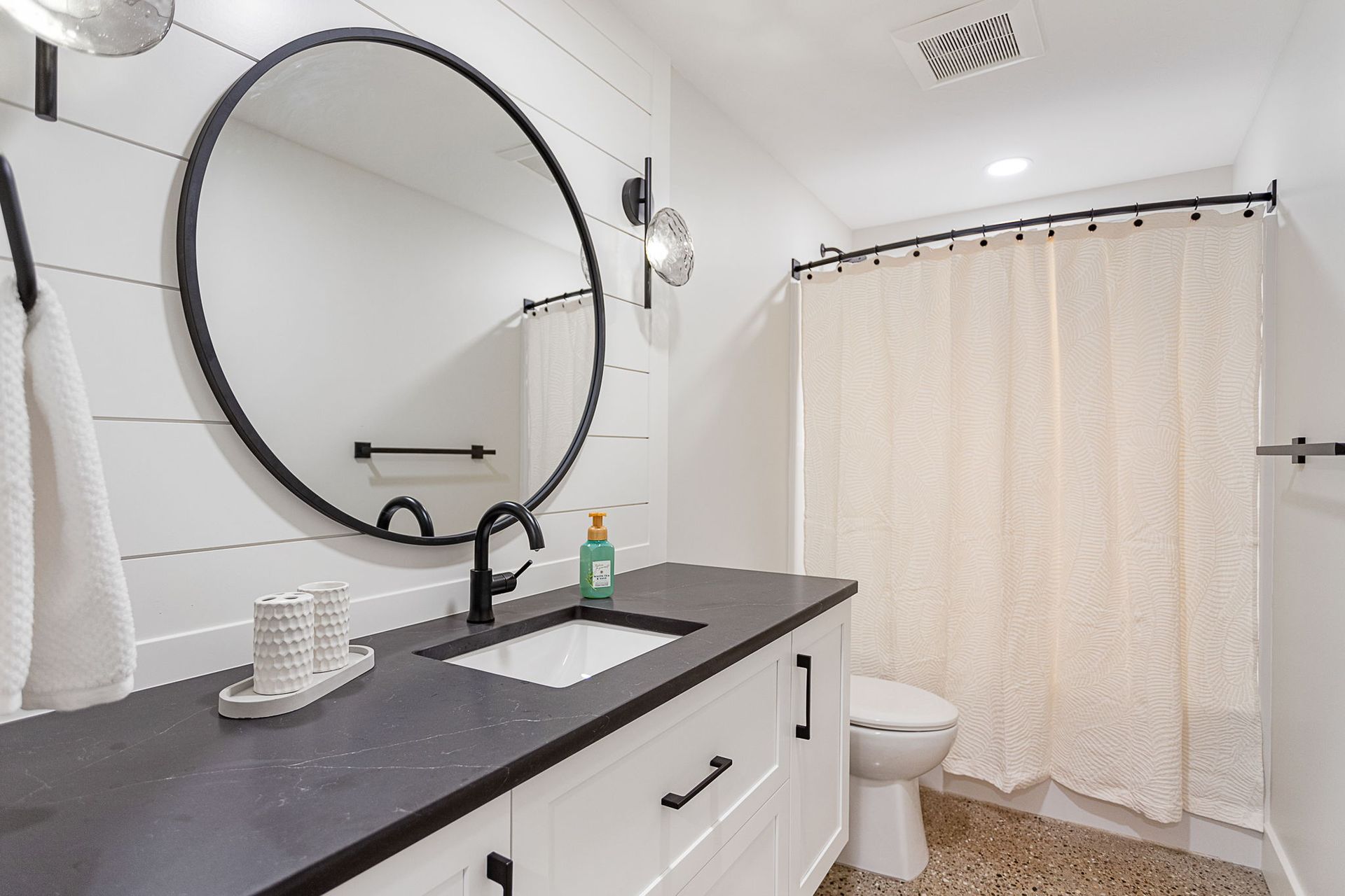 A modern bathroom featuring a white vanity with a dark countertop, a round black-framed mirror, and a shower curtain.