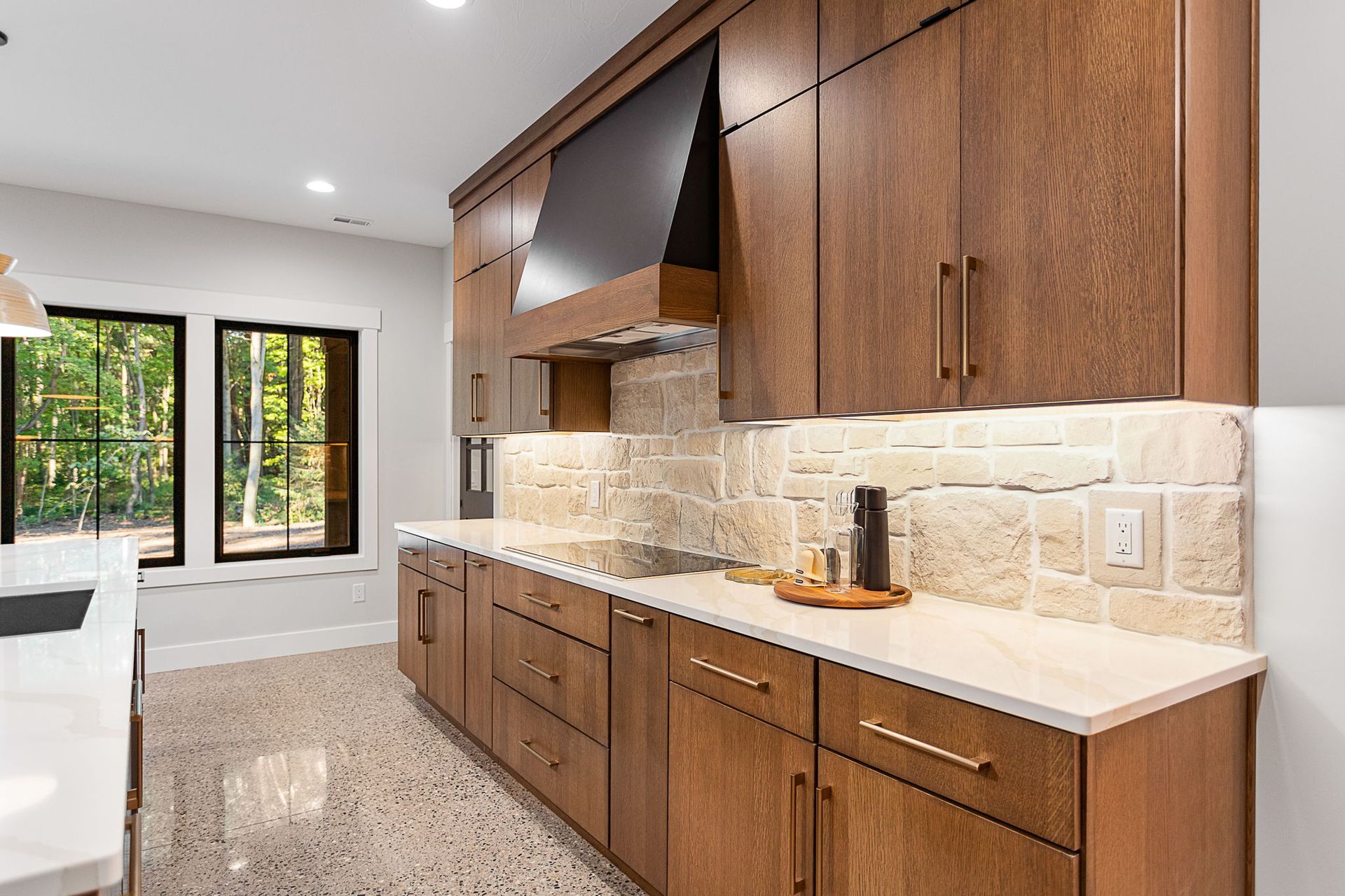 A modern kitchen with wood cabinets, stone backsplash, white countertops, and windows looking out onto a wooded area.