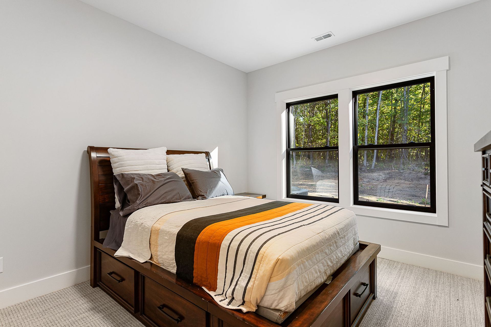 A bedroom with light gray walls, a wooden storage bed with a striped throw, and a window overlooking a forest.