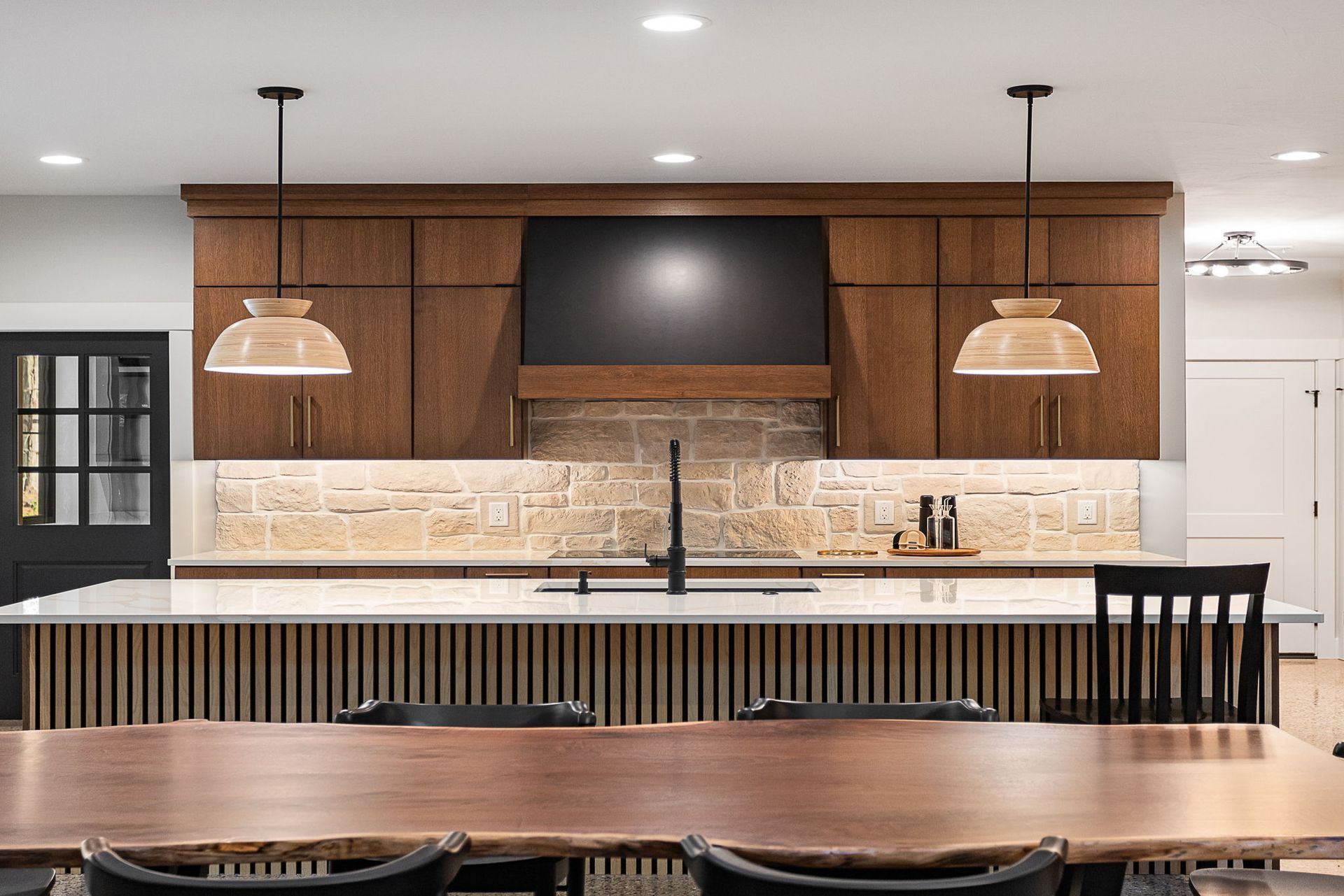 A modern kitchen with dark wood cabinets, stone backsplash, a large slatted island, and pendant lights over a wooden table.