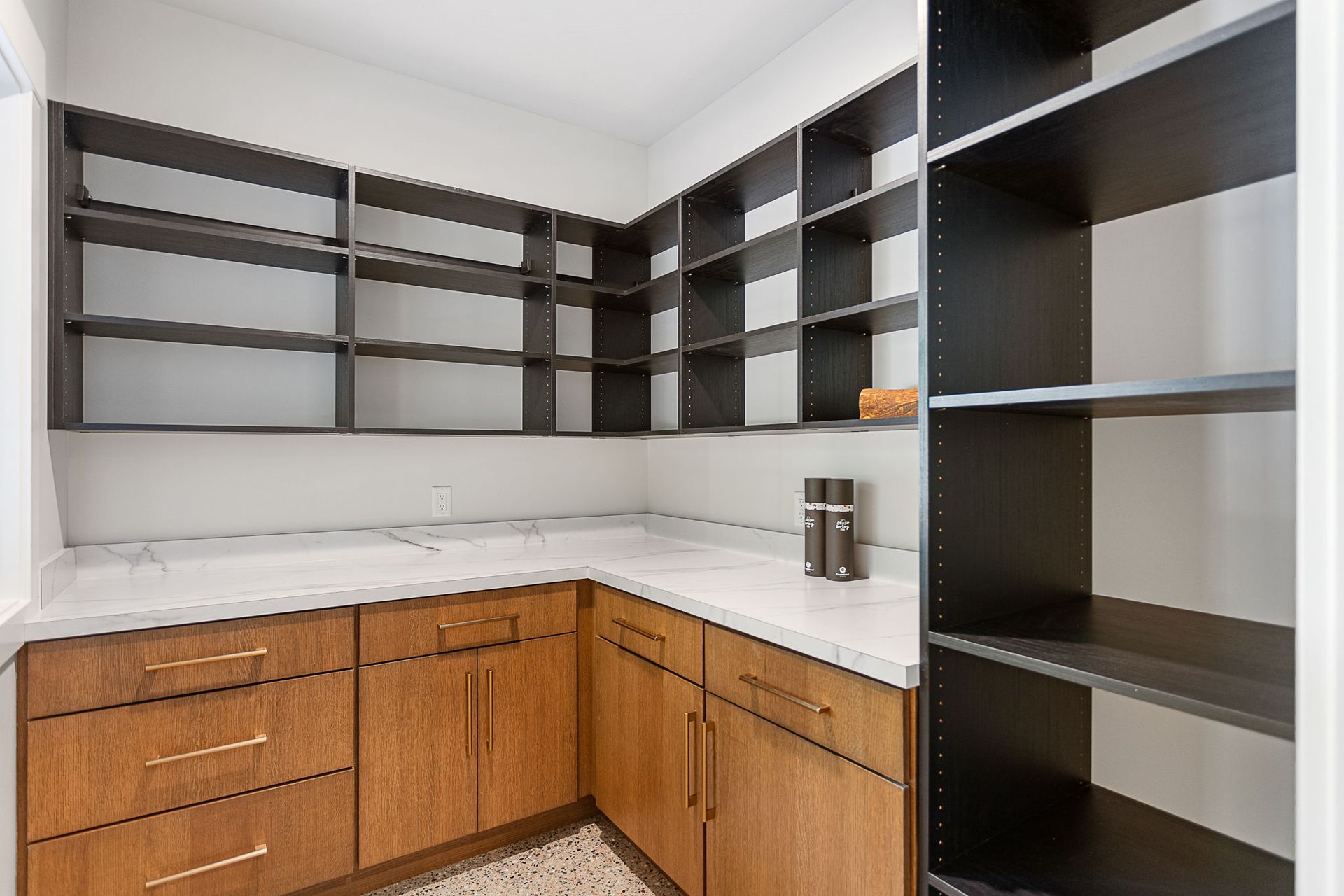 A corner pantry with dark wooden open shelves, light marble countertops, and light-stained wood cabinets with brass pulls.