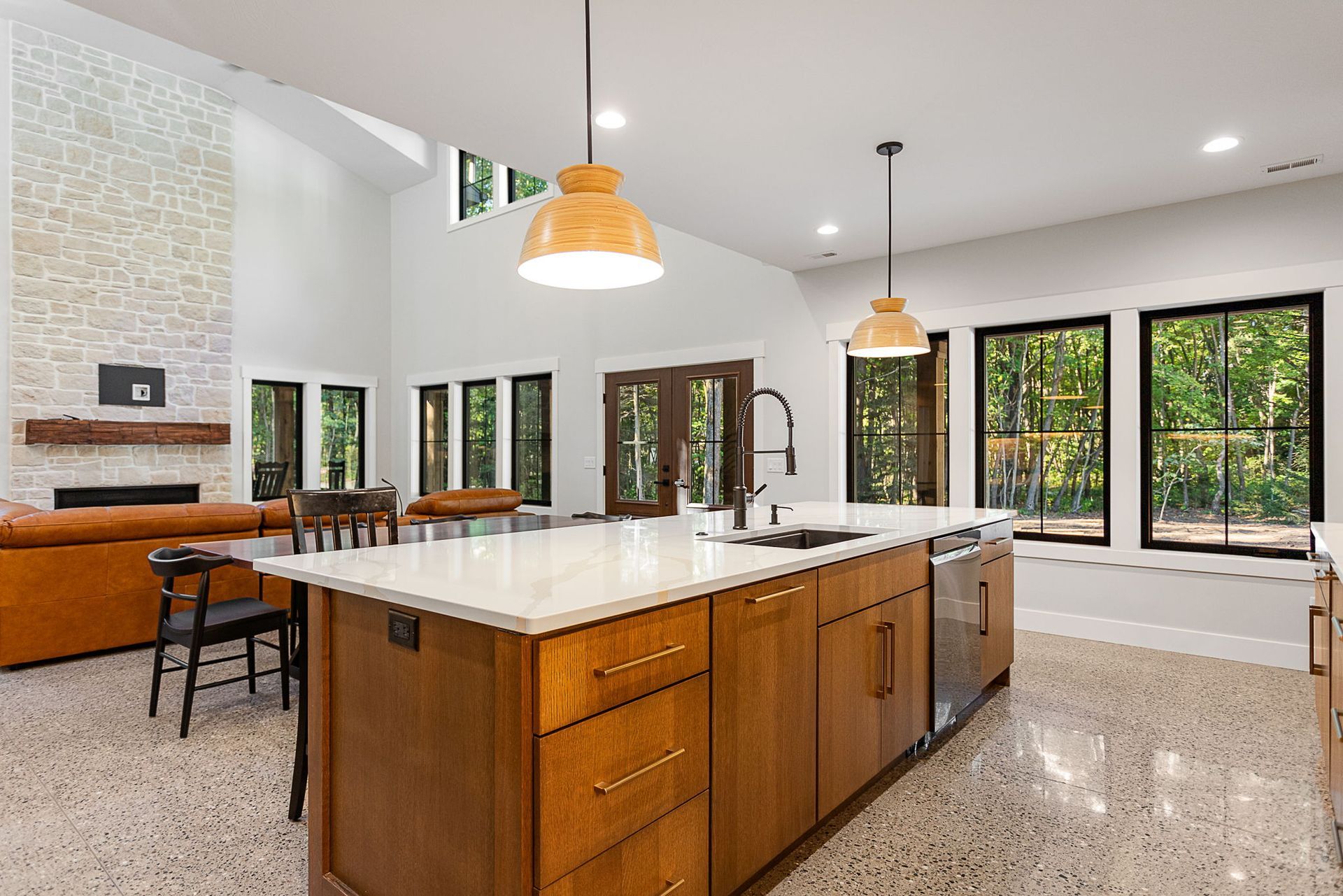 A modern kitchen with wood cabinets, a white island, pendant lights, and a stone fireplace in the background.