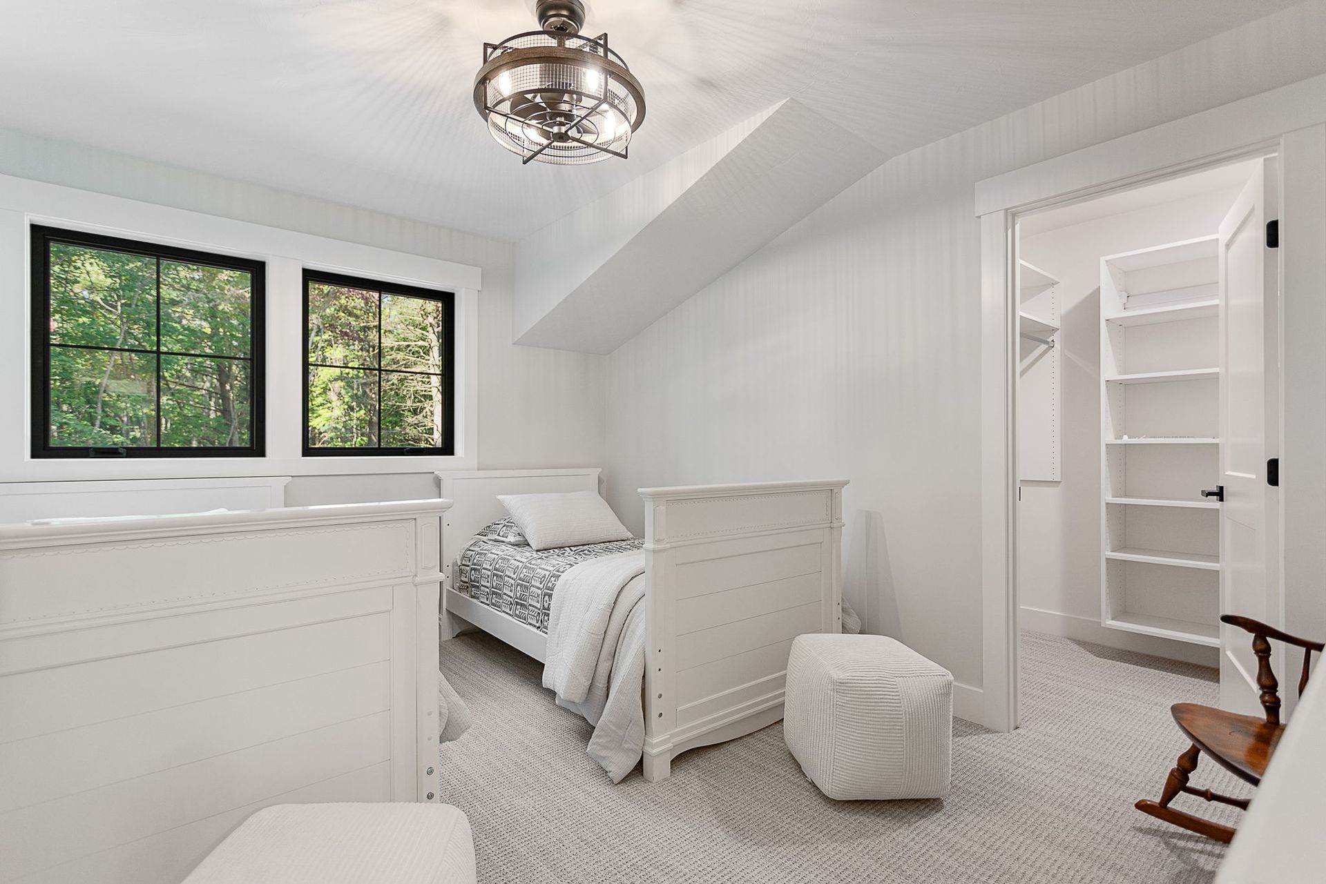 A bright bedroom featuring a single bed, a white ottoman, and a wooden rocking chair, with an open walk-in closet nearby.