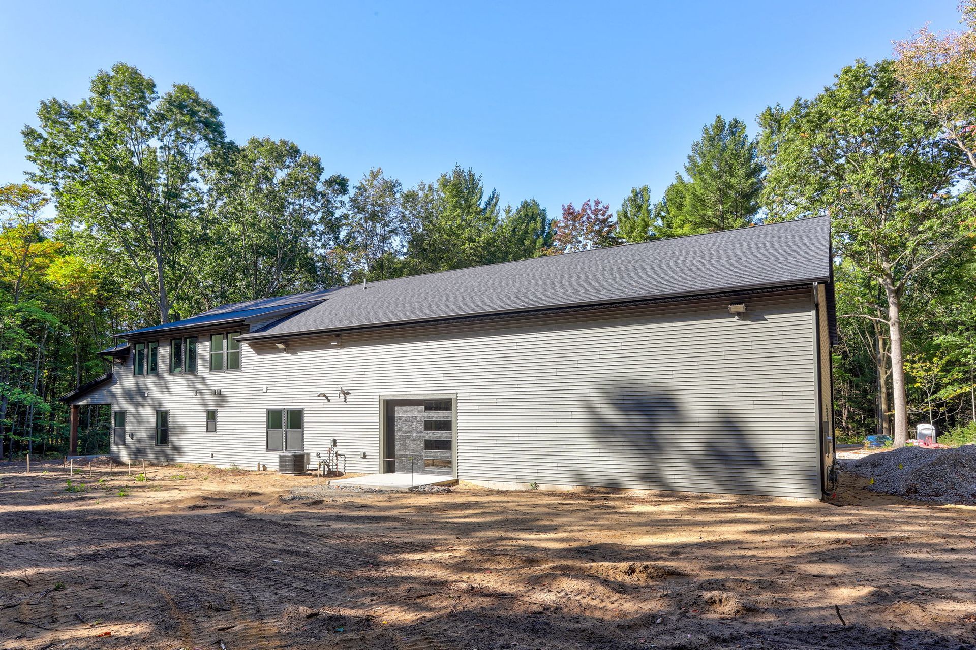 A modern, two-story house with gray horizontal siding and a dark roof, surrounded by trees on a cleared dirt lot.