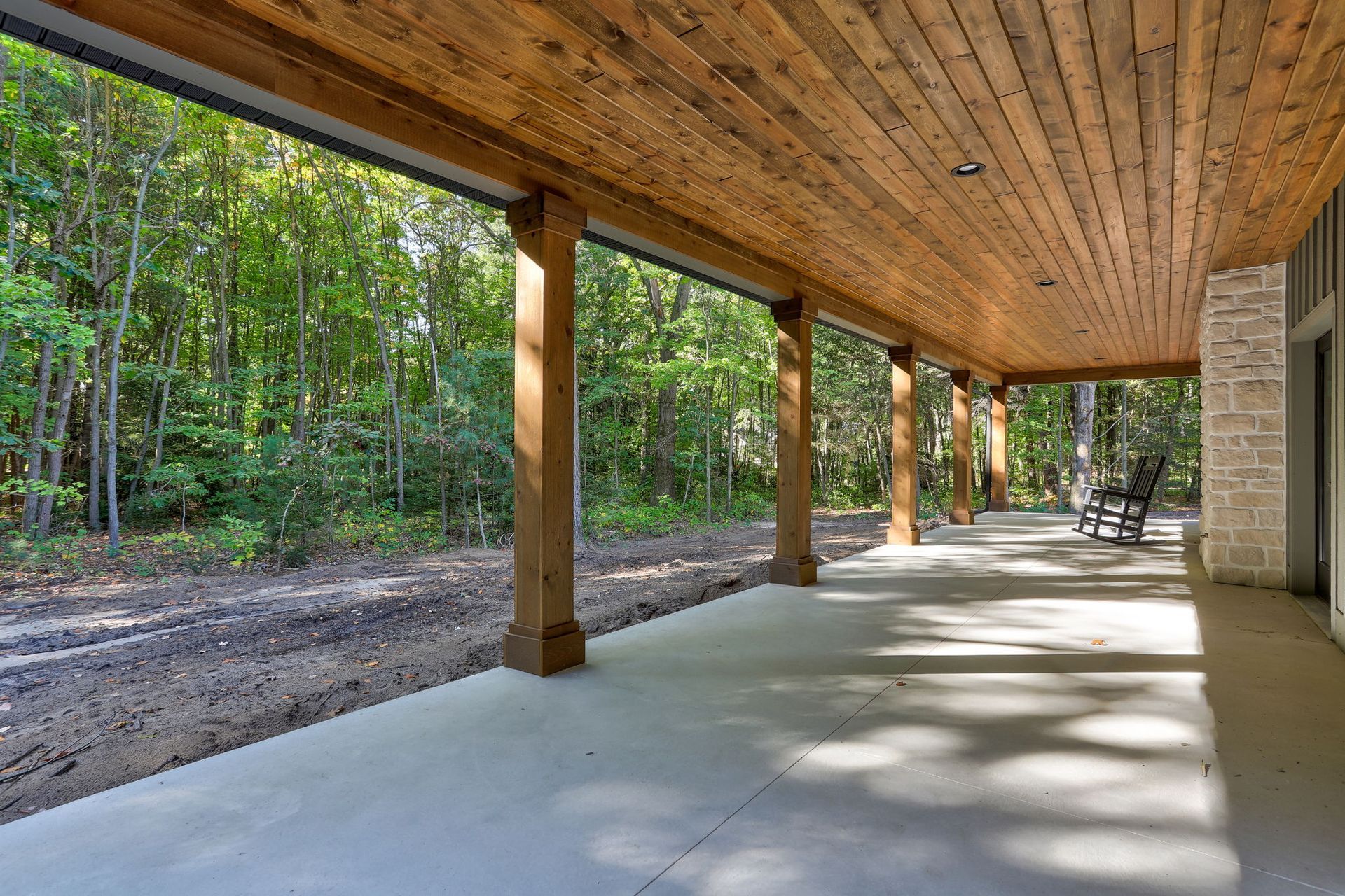 A wide, covered porch with wood-paneled ceilings, concrete floors, and wooden pillars overlooking a wooded area.