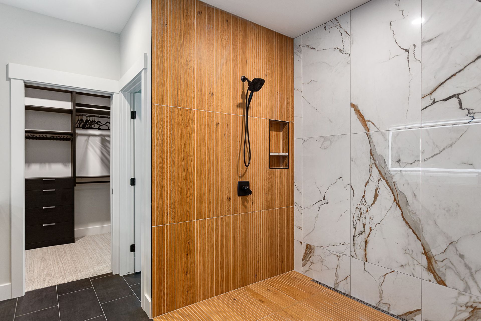 A shower featuring a wood-slat wall and floor alongside large white marble-patterned tiles, with a walk-in closet nearby.