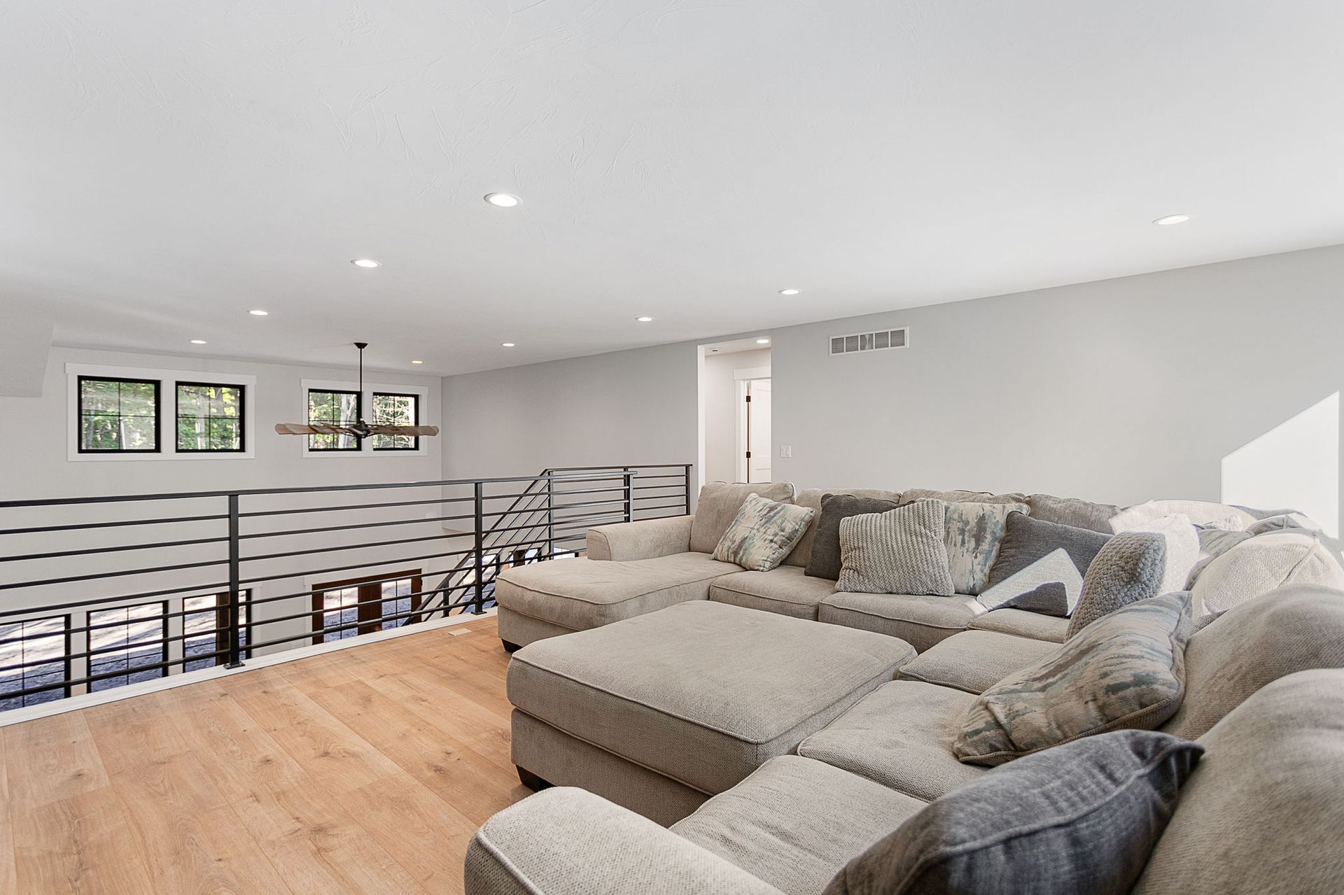 A large, beige sectional sofa sits in a bright, open loft area with light wood floors and a horizontal metal railing.