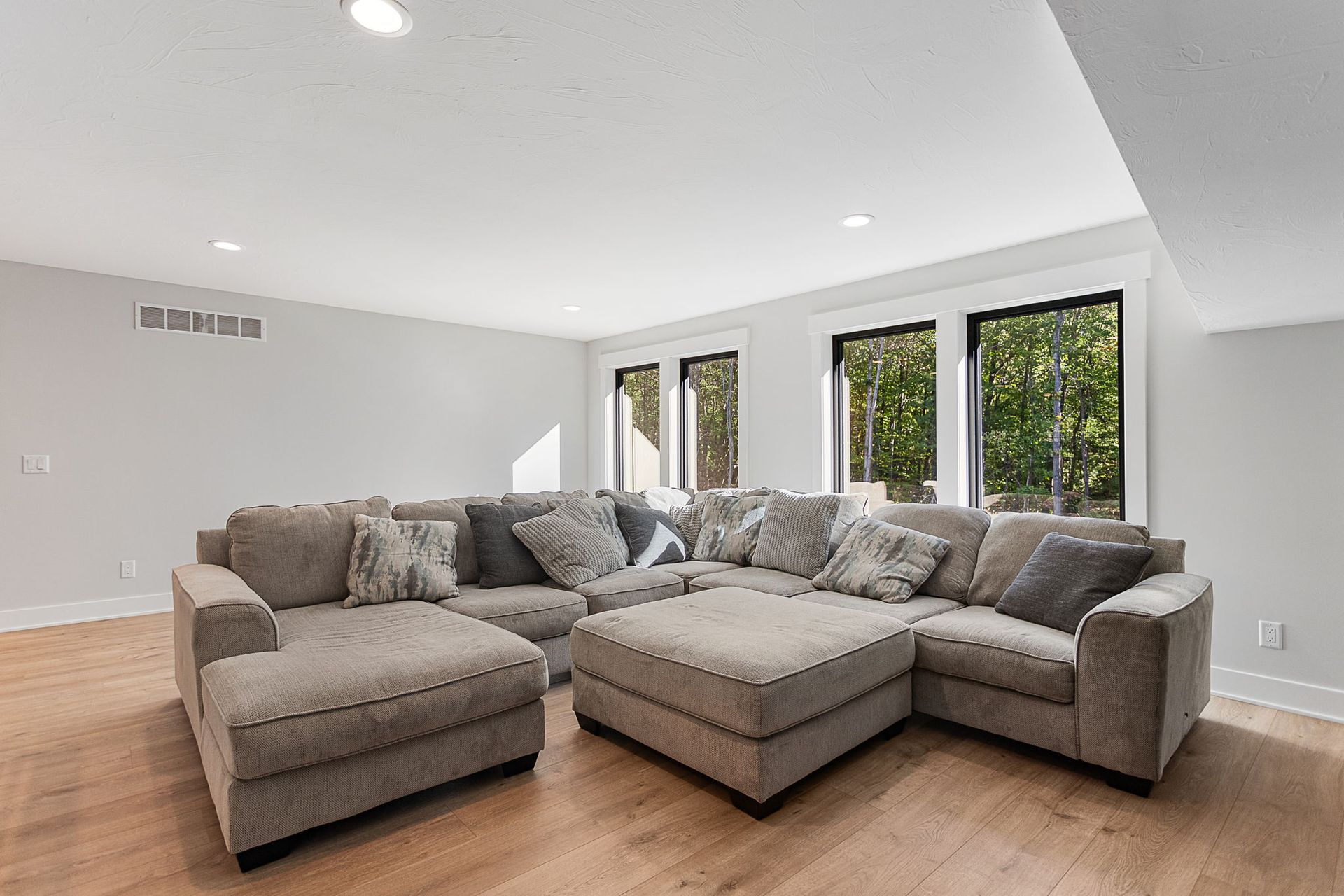 A large gray sectional sofa with an ottoman sits in a sunlit room with hardwood floors and windows looking out to trees.