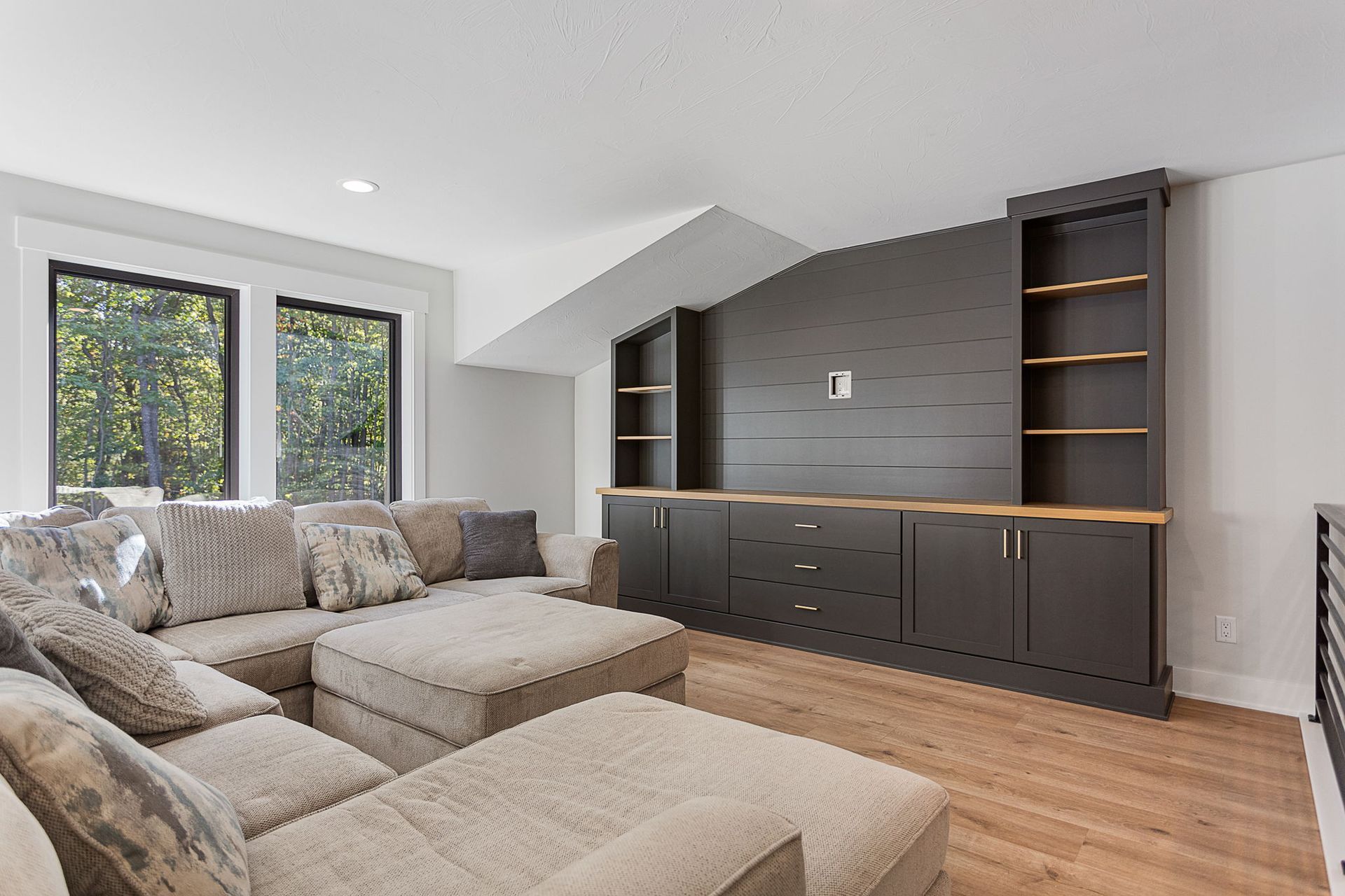 A light-filled living room with a large, grey sectional sofa facing a dark-grey media console with built-in shelves.