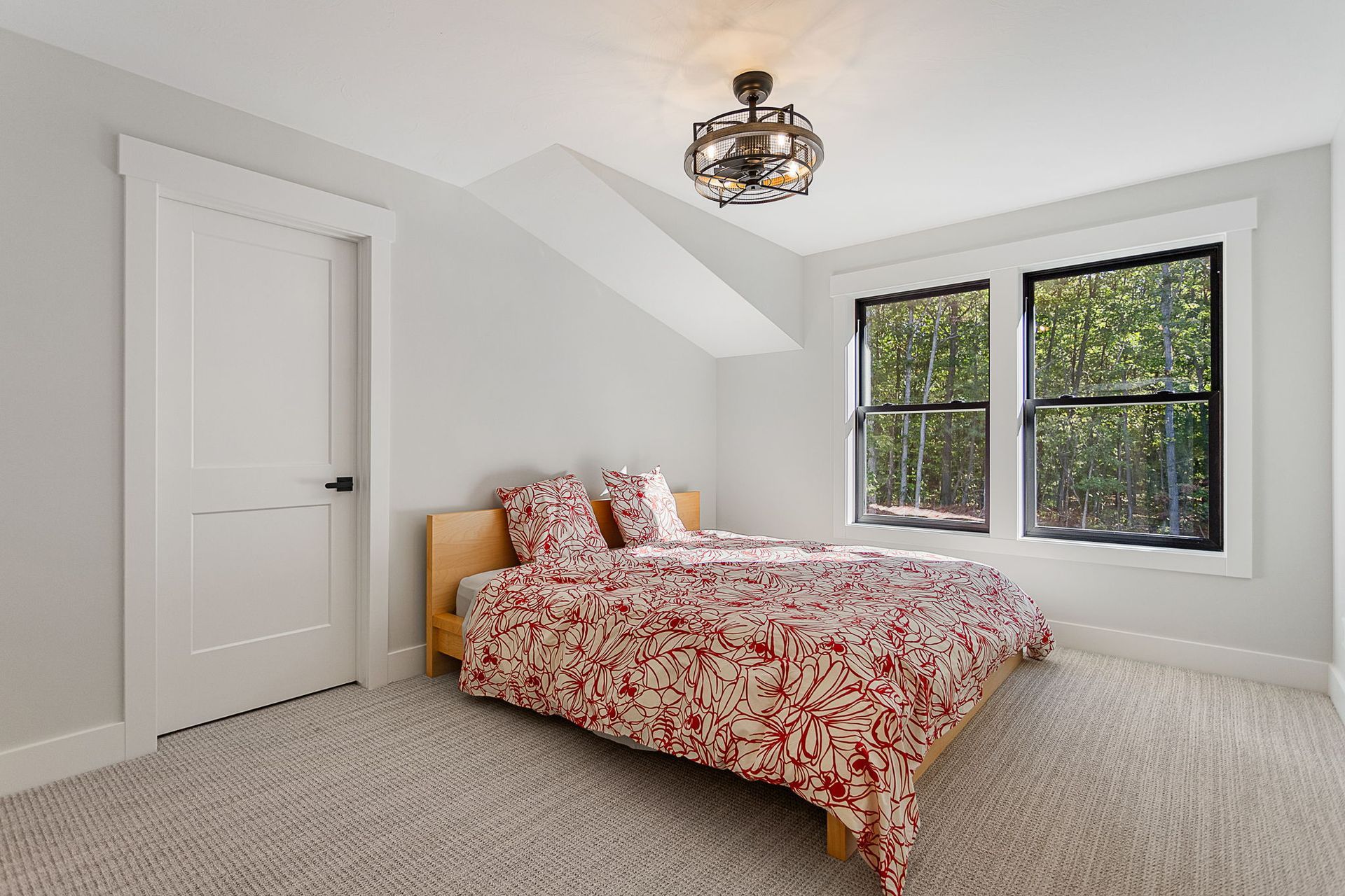 A bedroom with a bed featuring a red and white patterned comforter, light grey walls, carpet, and a large window.