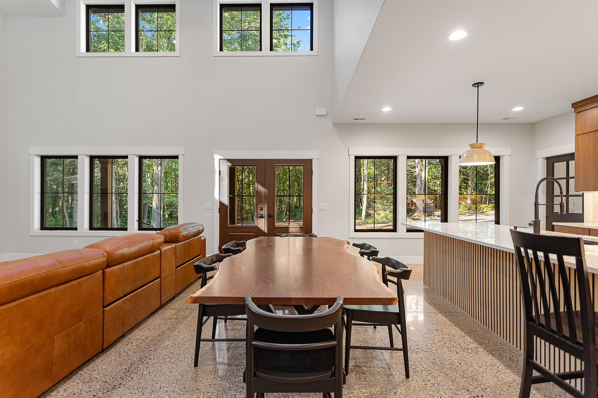 A spacious, open-concept room featuring a long wooden dining table, a tan leather sectional sofa, and kitchen island.