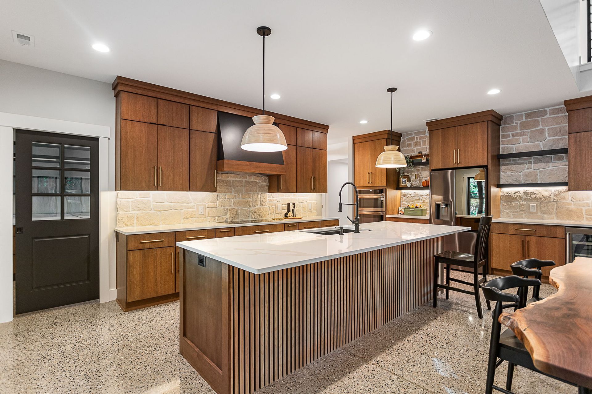 A modern kitchen featuring wooden cabinets, a large island with a slatted base, stone backsplashes, and terrazzo floors.