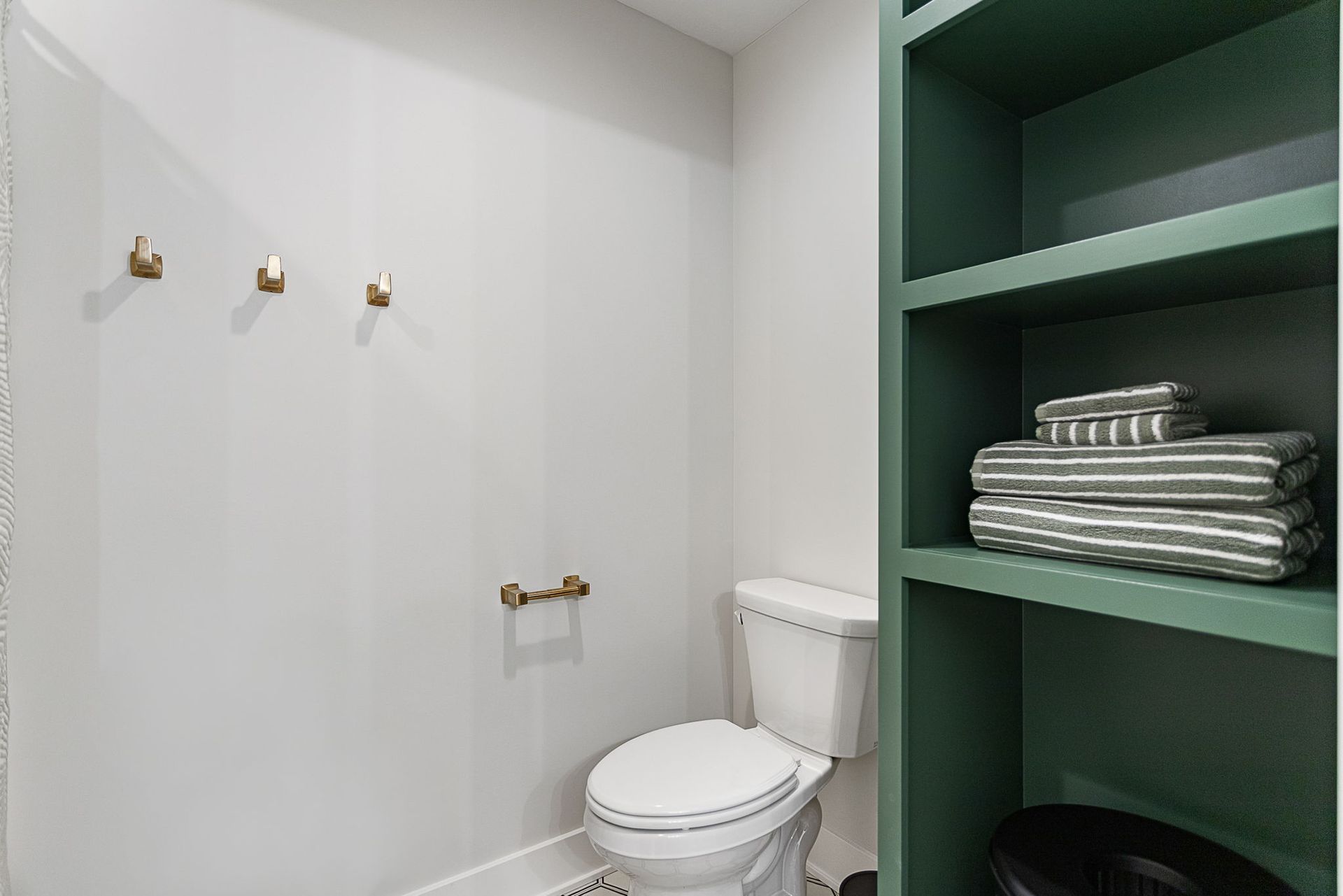 A bathroom featuring a toilet, three gold wall hooks, and a built-in green shelf holding stacked grey striped towels.