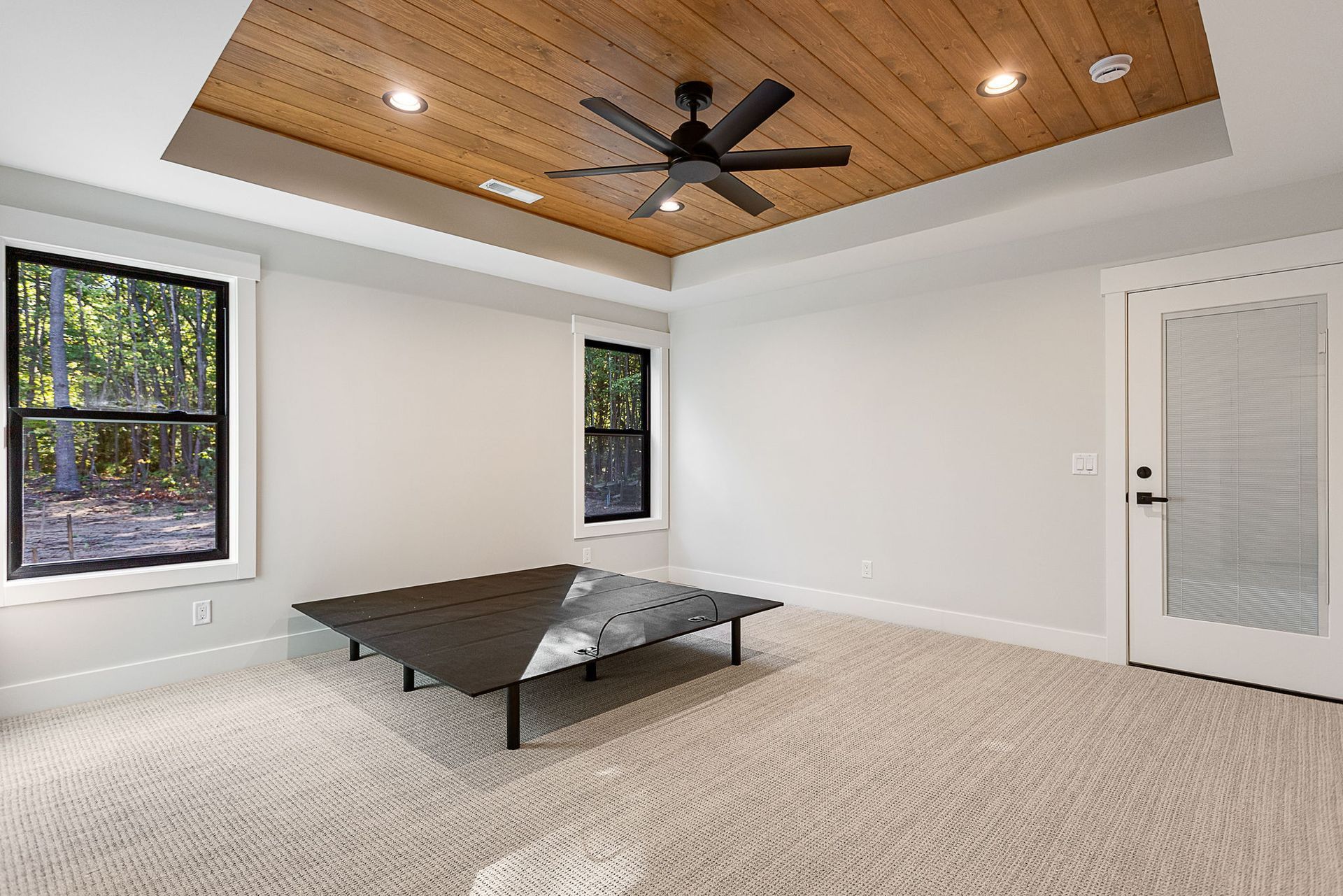 A bright, modern bedroom featuring a wood-plank tray ceiling, a ceiling fan, two windows, and a black platform bed frame.