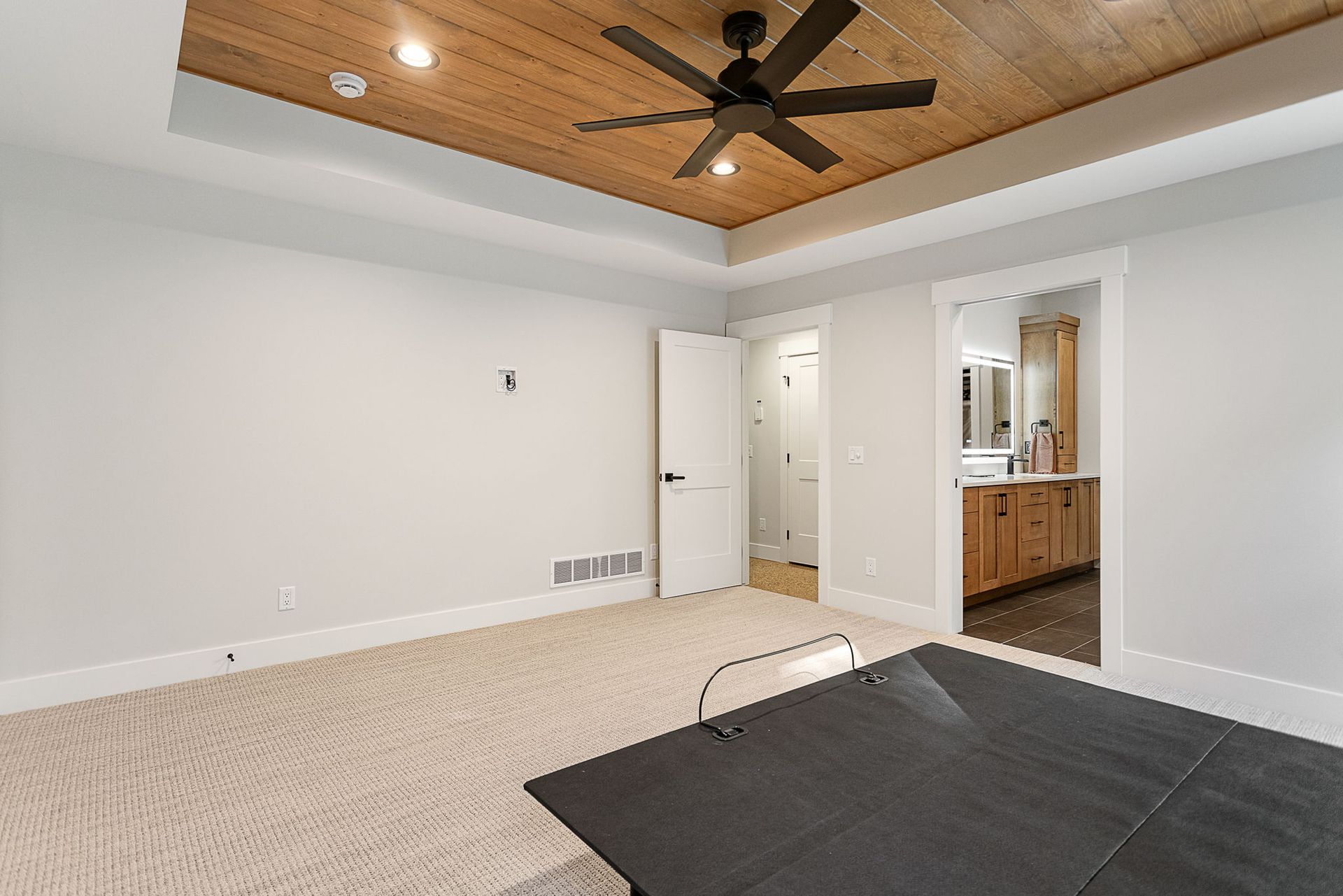 A bedroom with beige carpet, a white door, and a wooden tray ceiling with a fan, looking into an adjacent bathroom.