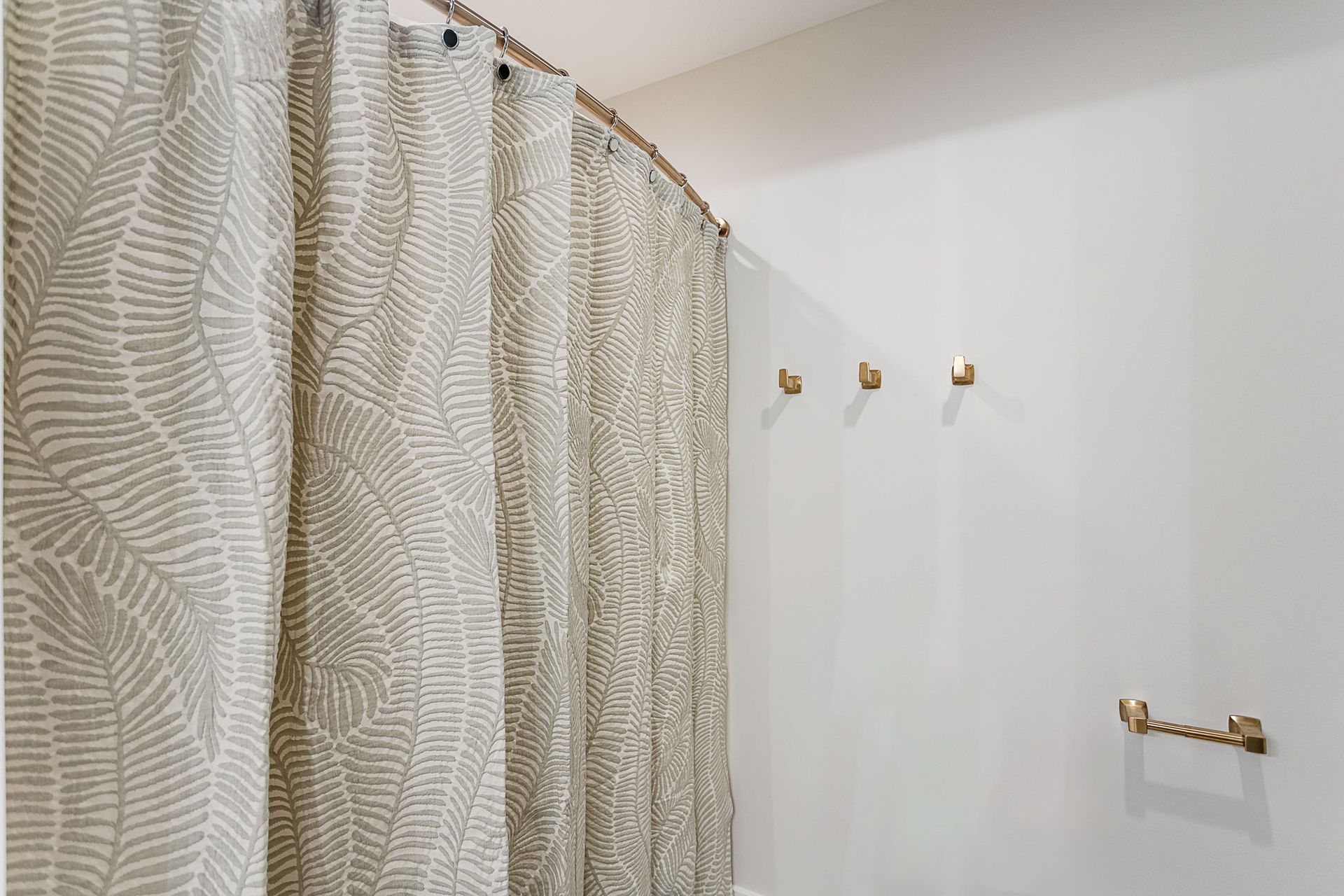 A shower curtain with a beige fern pattern hangs on a curved rod, near three small gold wall hooks and a gold towel bar.