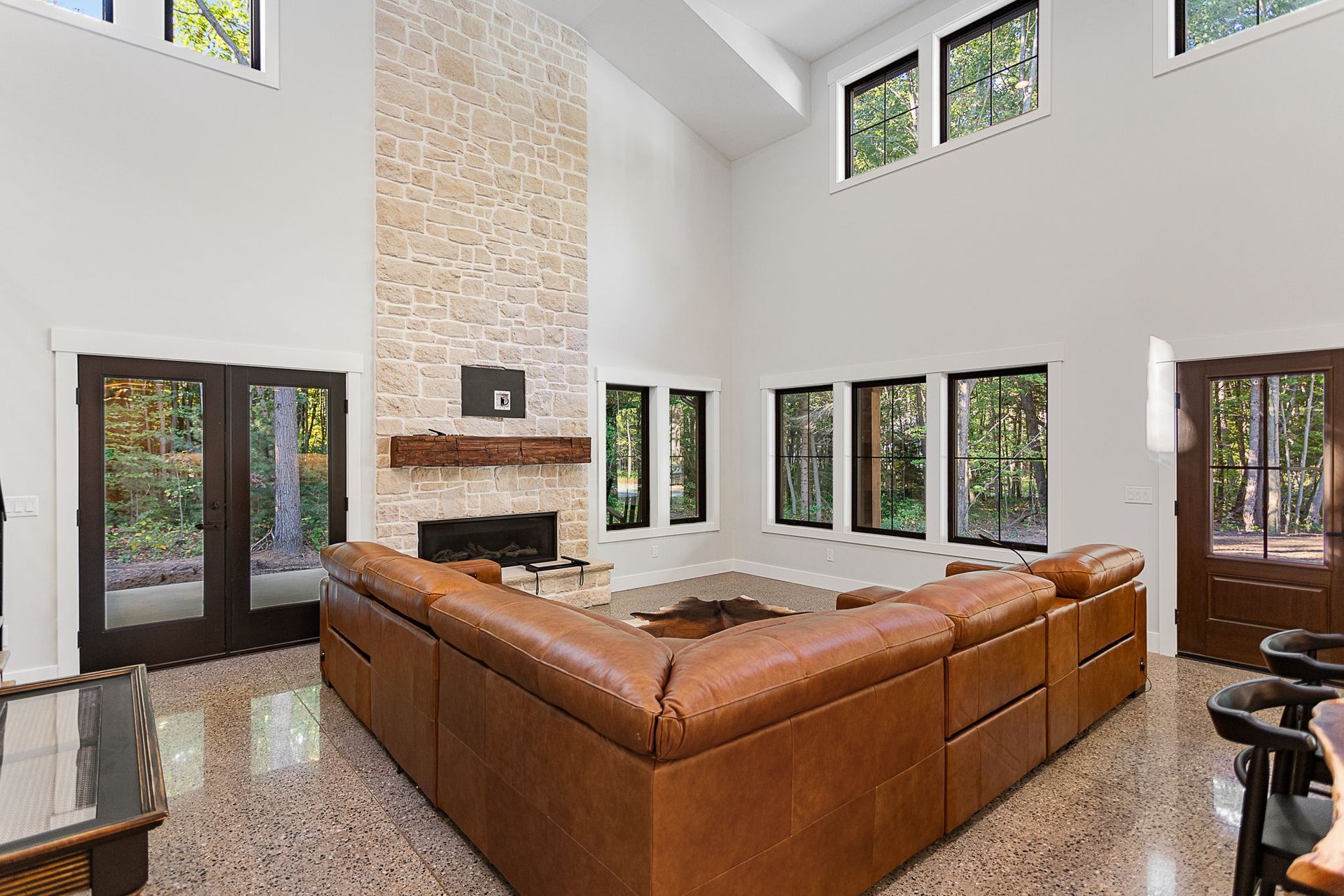 A living room with a large tan leather sectional, a tall stone fireplace, glossy floors, and multiple large windows.