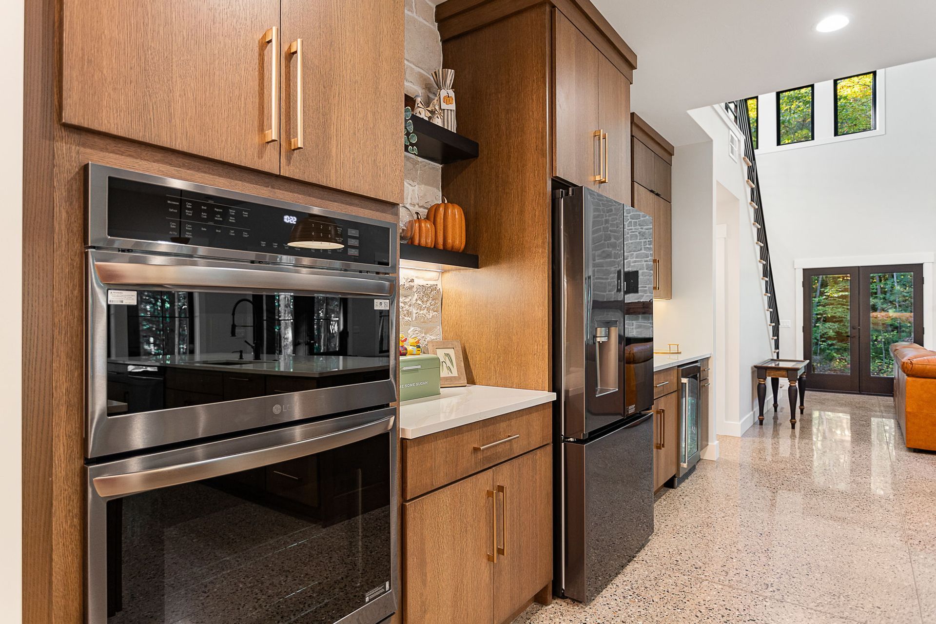 Modern kitchen with wood cabinets, stainless steel double wall oven, and dark refrigerator, opening to a sunlit room.
