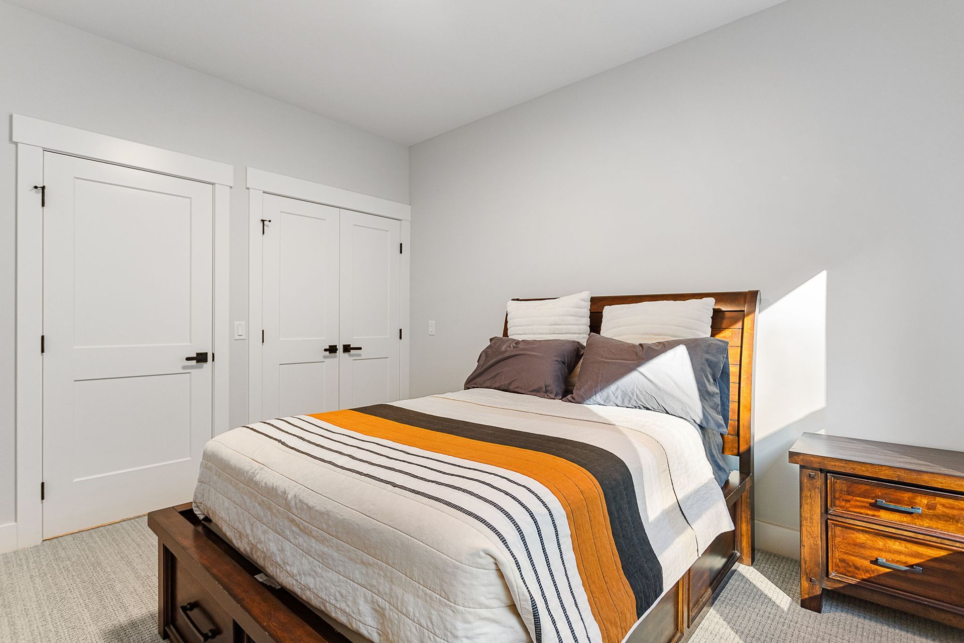 A bedroom with a wooden bed frame, a striped comforter in orange, gray, and white, and two white closet doors.