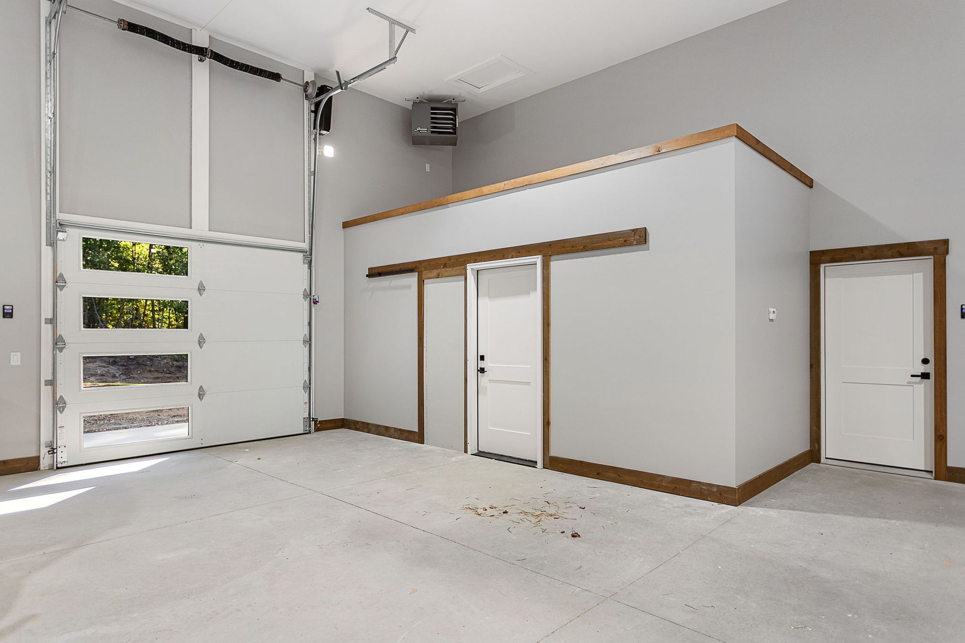 A modern, empty garage with white walls, a roll-up glass panel door, two interior doors, and wood-toned trim.