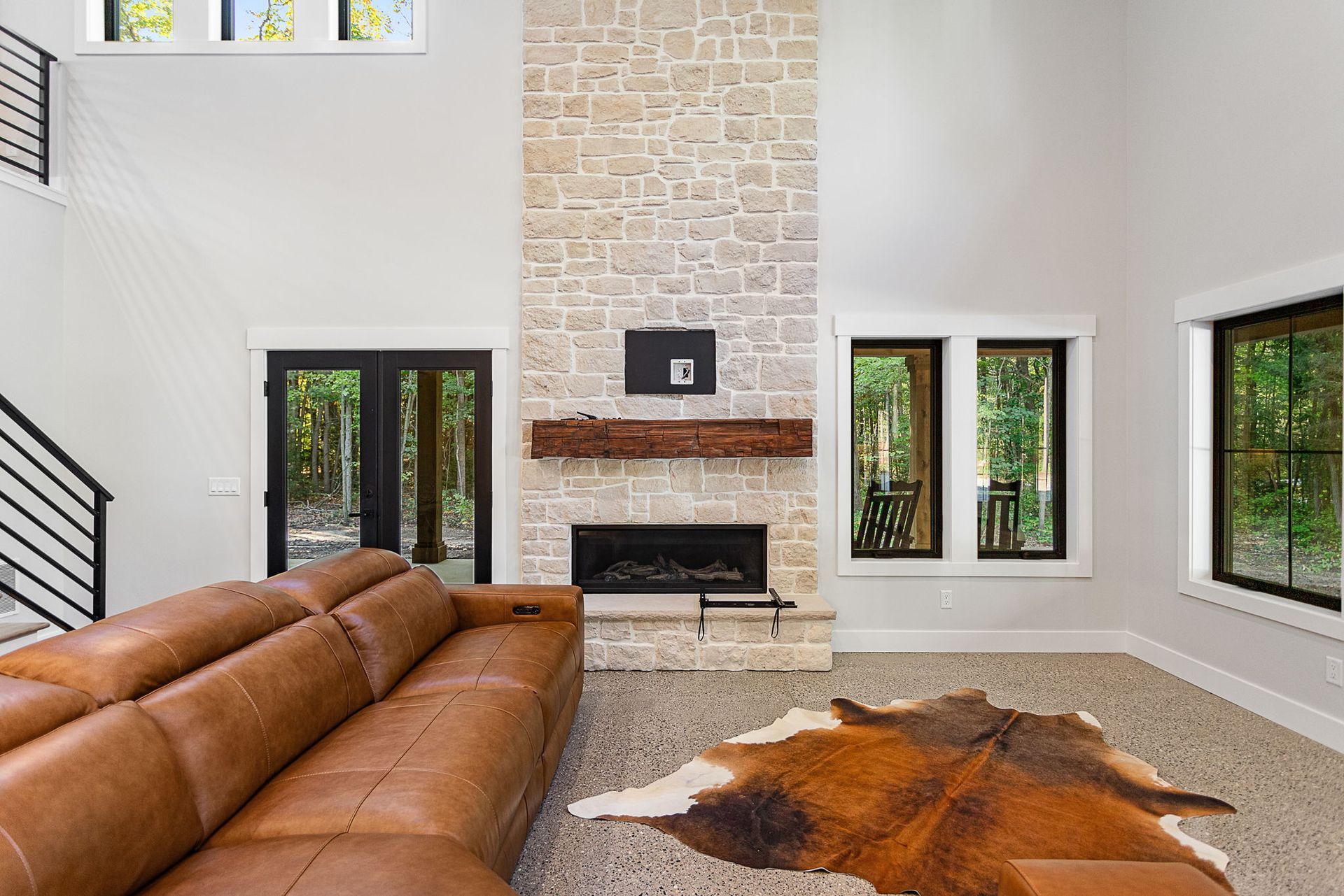 Modern living room with a leather sectional, stone fireplace, and cowhide rug under high ceilings with large windows.