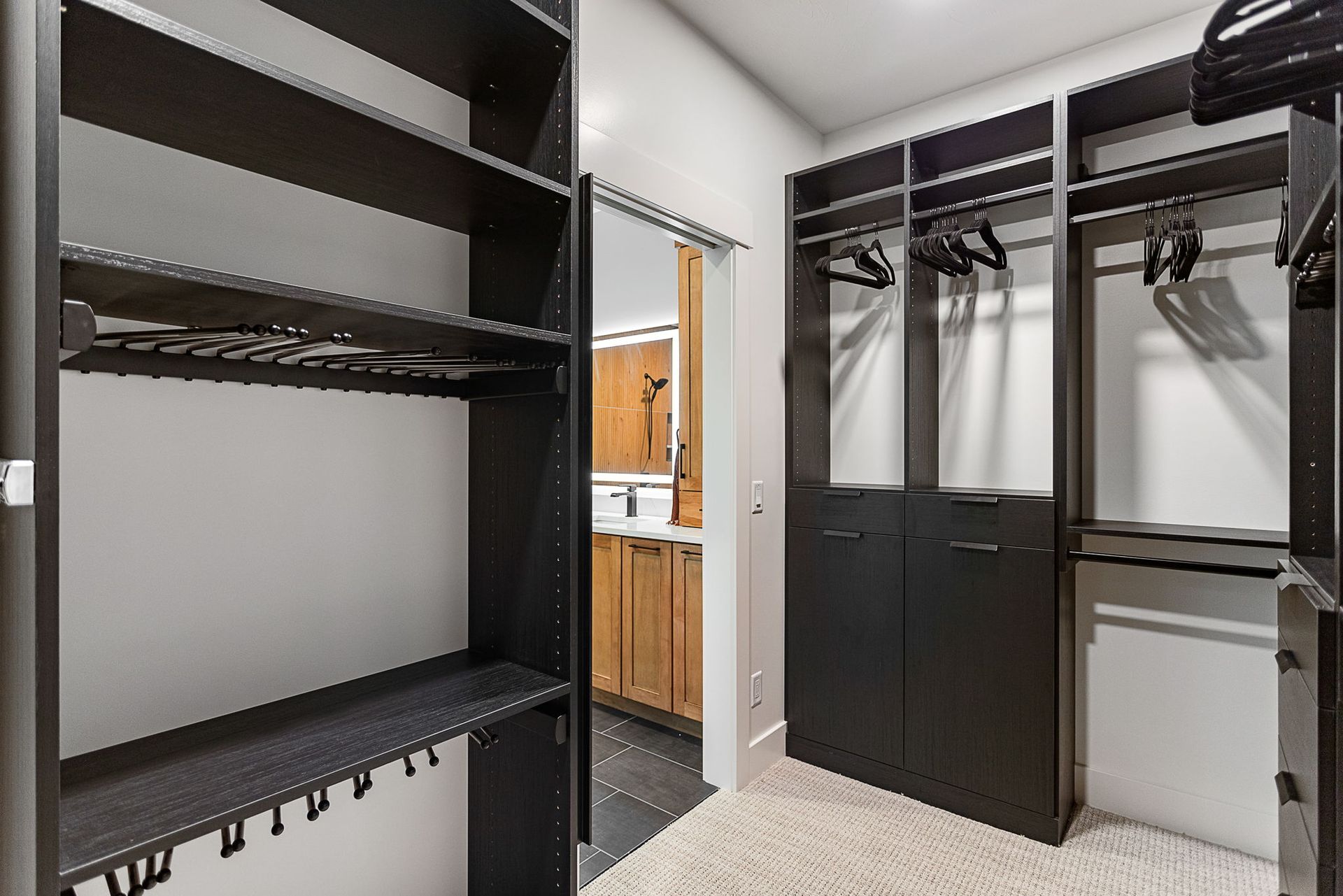 A walk-in closet featuring dark wood shelving, hanging rods, and carpeted floors, leading into a bathroom.