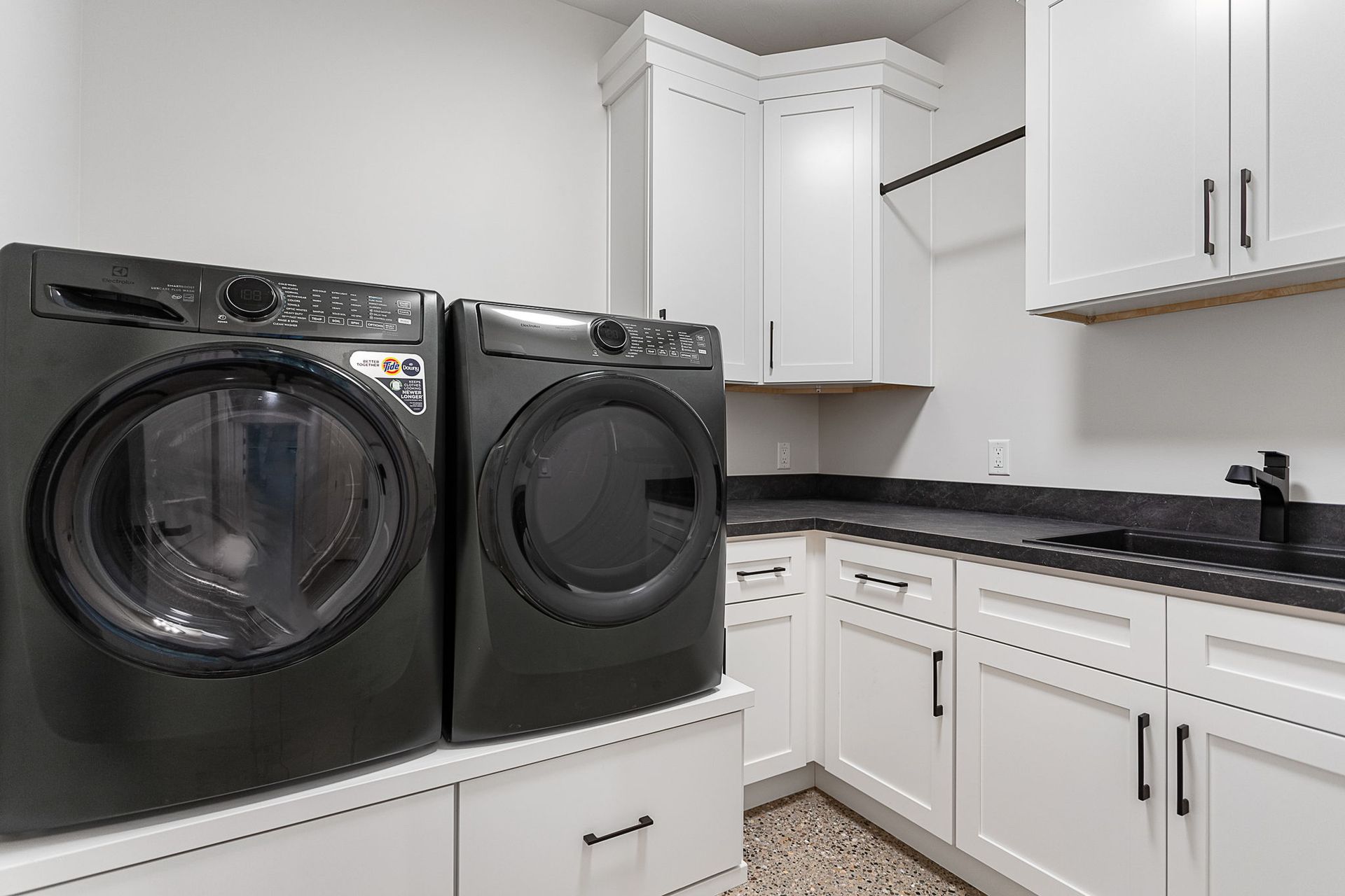 A laundry room with dark grey washer and dryer units on white pedestals, next to white cabinets and a dark countertop.