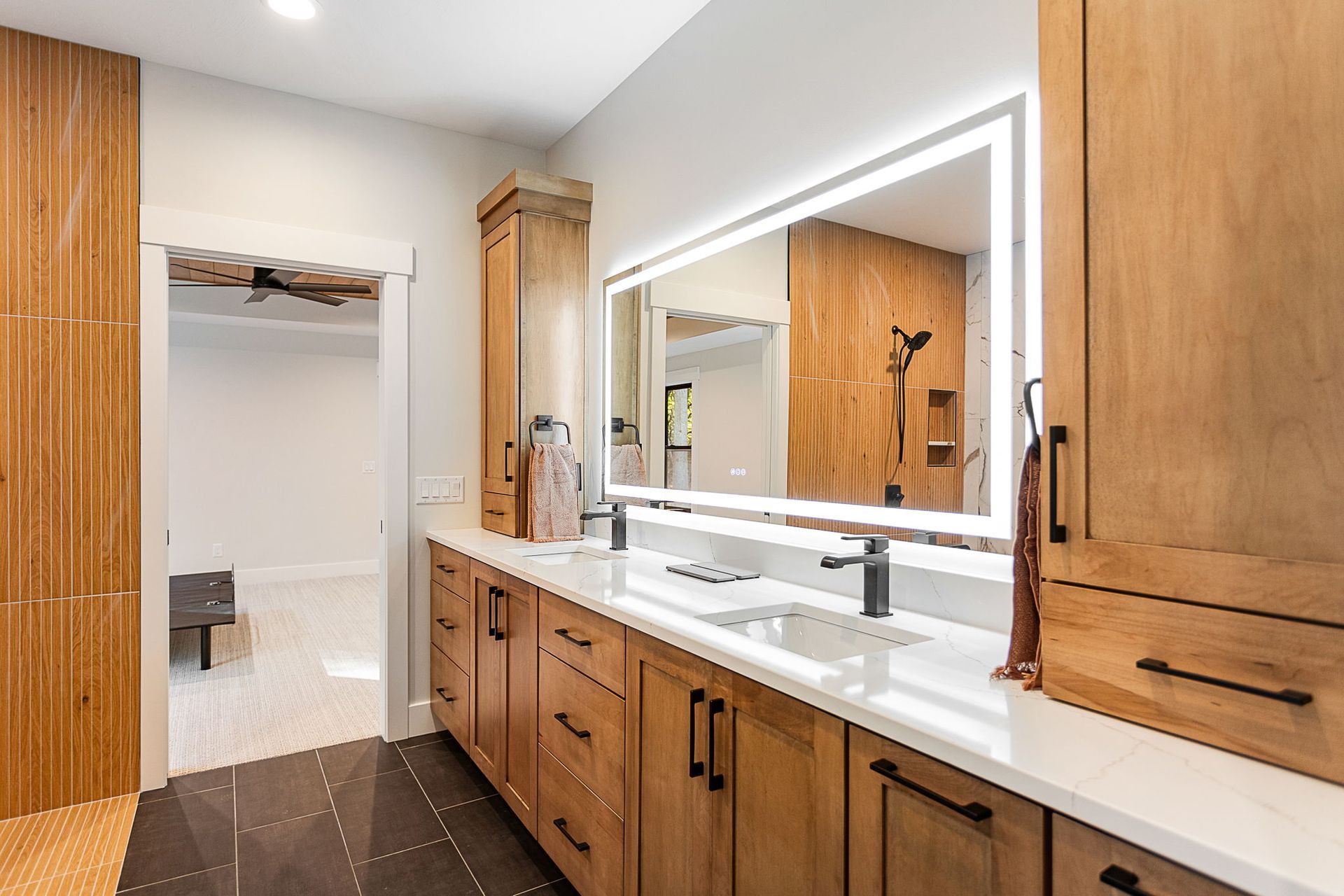 Modern bathroom featuring warm wood cabinetry, a double vanity with white countertops, and a large backlit mirror.