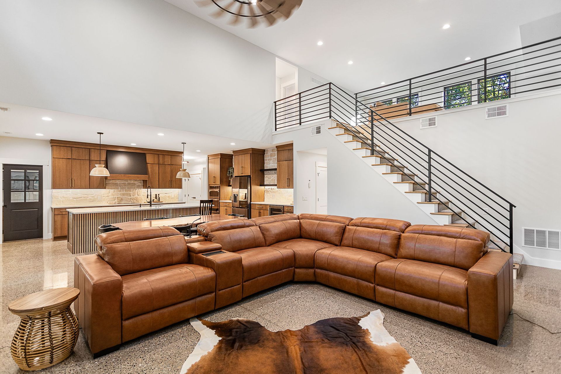 A modern open-plan living room with a brown leather sectional, kitchen, and staircase with a metal railing.