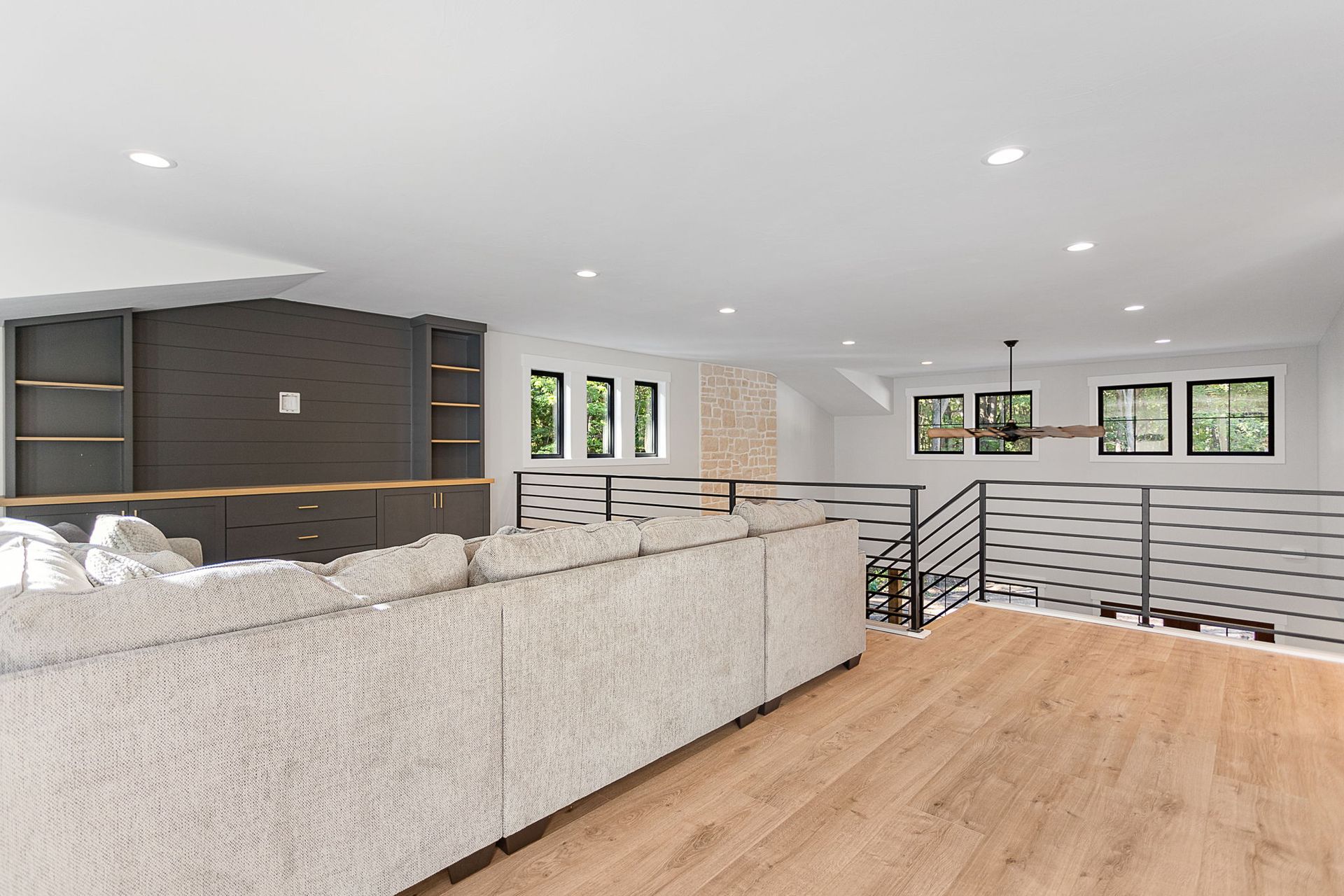 A modern living room with light wood floors, a large grey sectional, dark wood built-in shelving, and a metal stair railing.