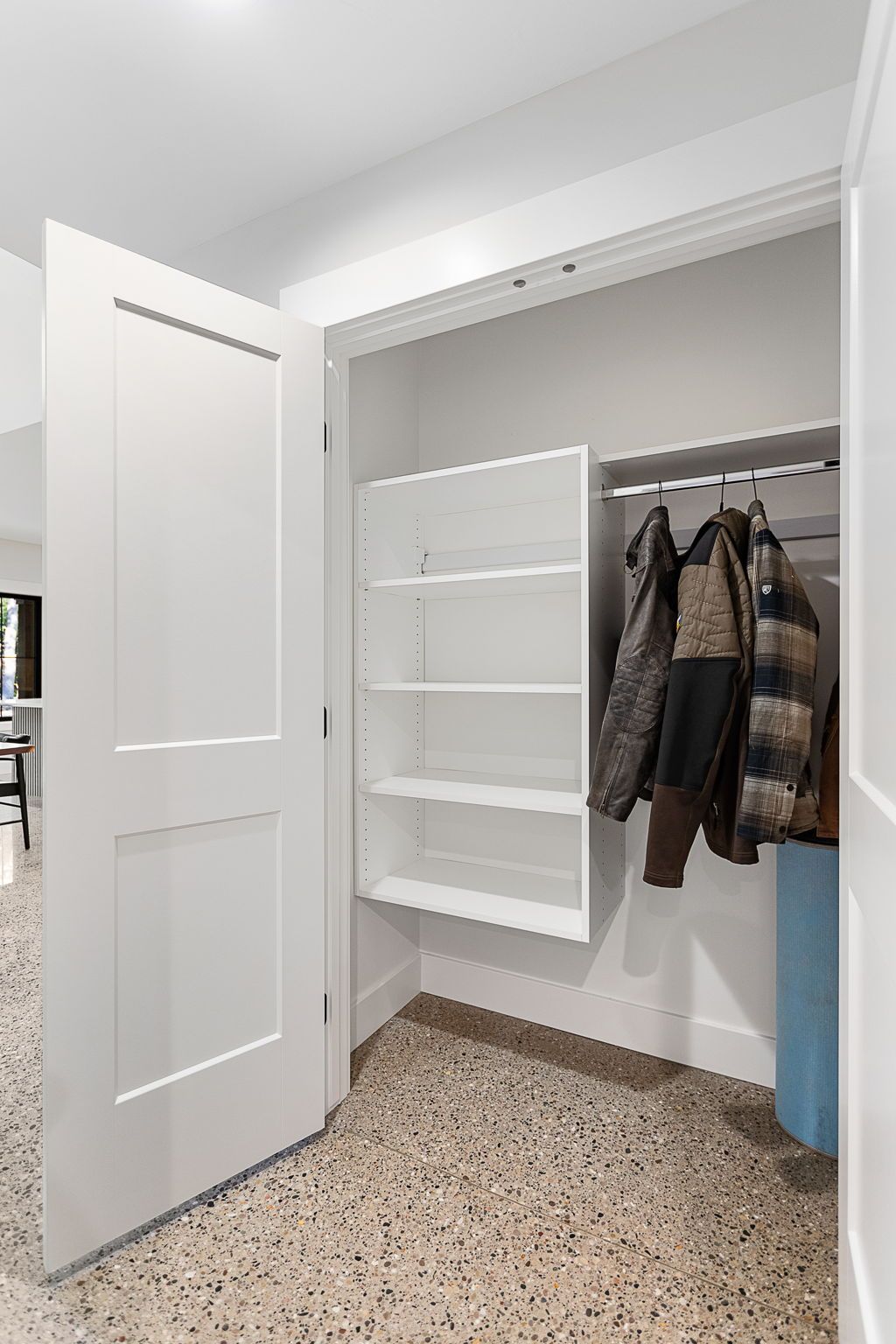 A closet with white shelves mounted on the left and a rod holding several hanging jackets on the right, over terrazzo floors.