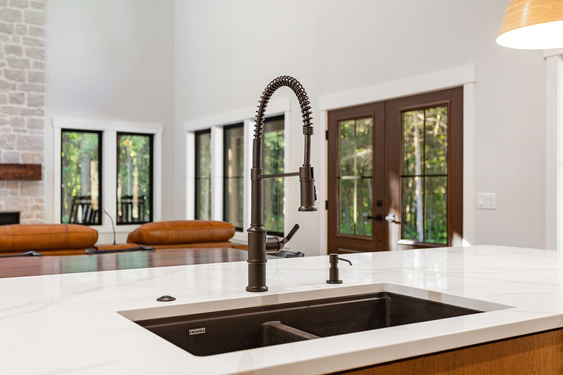 A bronze pull-down kitchen faucet and dual-basin sink set in a white countertop with a living area in the background.