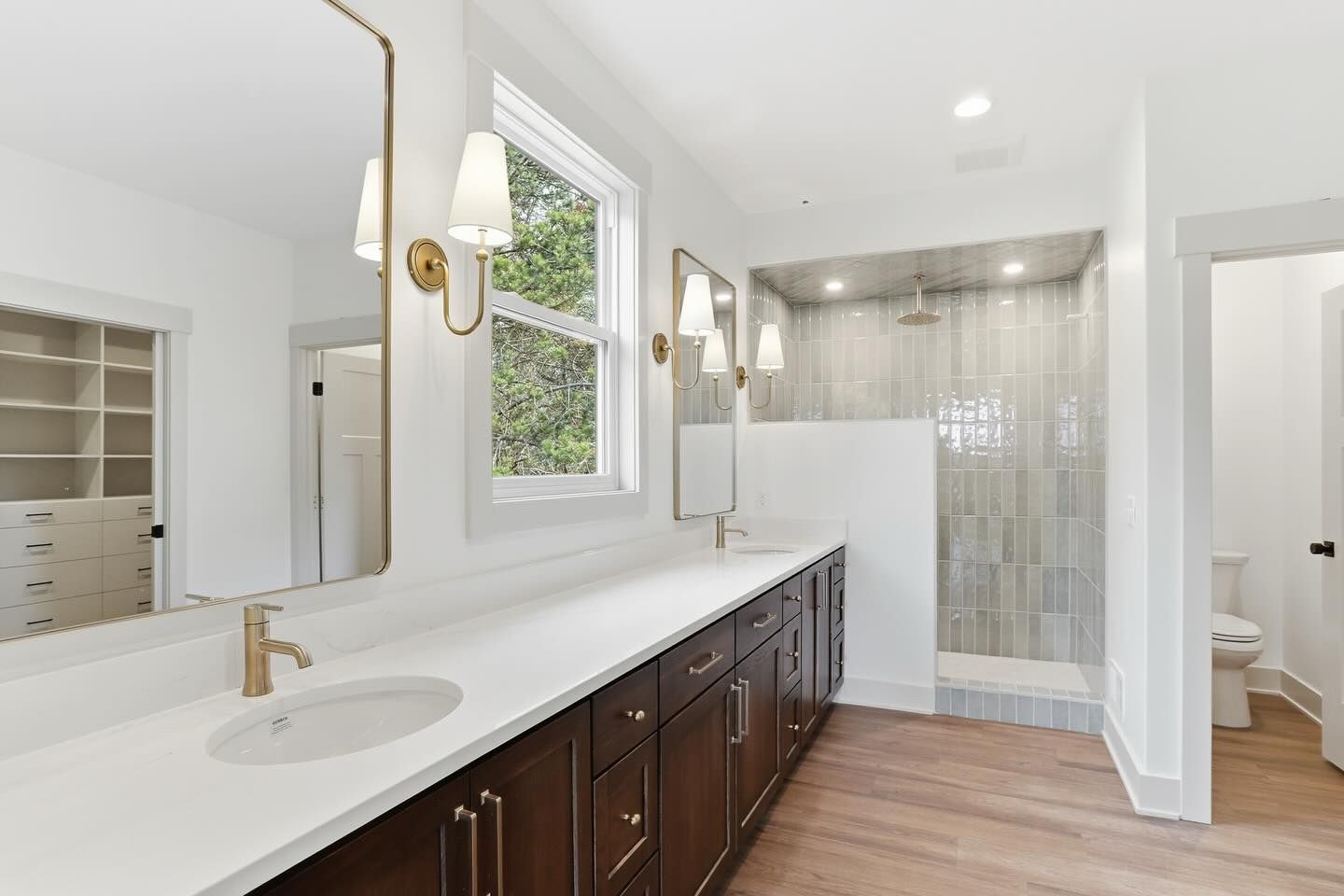 A bright, modern bathroom featuring a white vanity with gold faucets, dark wood cabinets, a glass shower, & a walk-in closet.