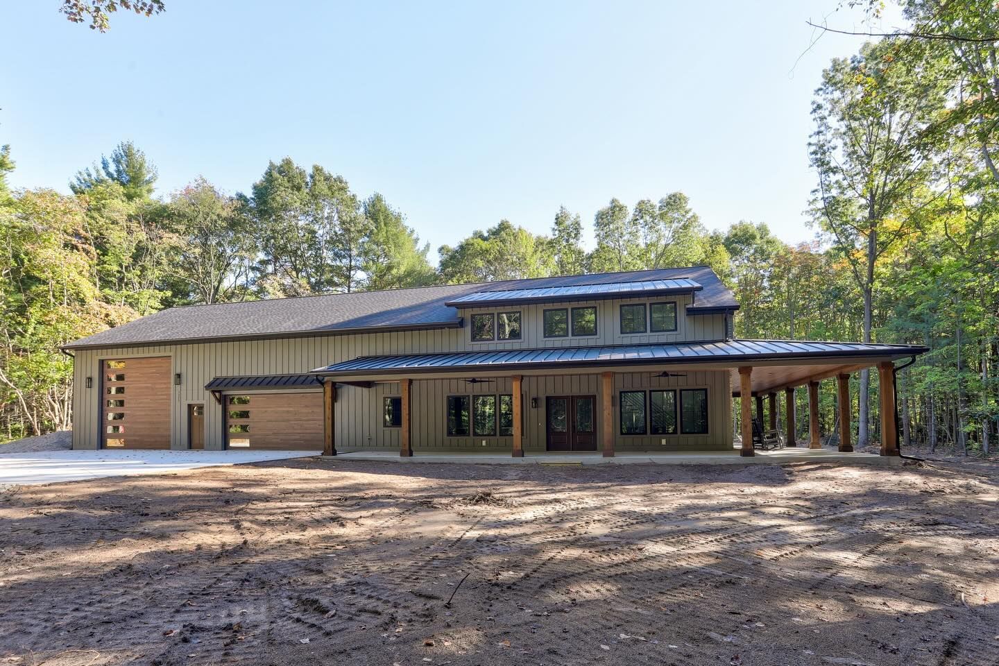 A tan, modern farmhouse with a dark metal roof and wooden porch pillars sits on a wooded lot with a freshly cleared yard.